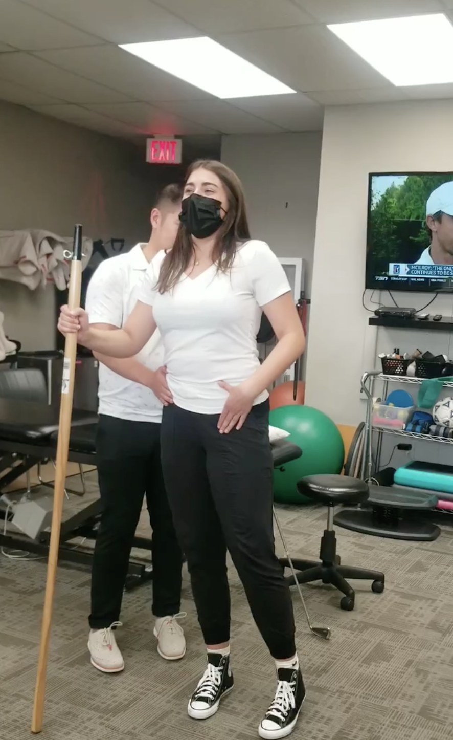 Physiotherapist, Jia Chen, is standing inside the Physiotherapy room at Mainway Physiotherapy teaching a client how to do a hip abduction. The female client is standing and holding a pole for balance with her leg slightly kicked to the side.
