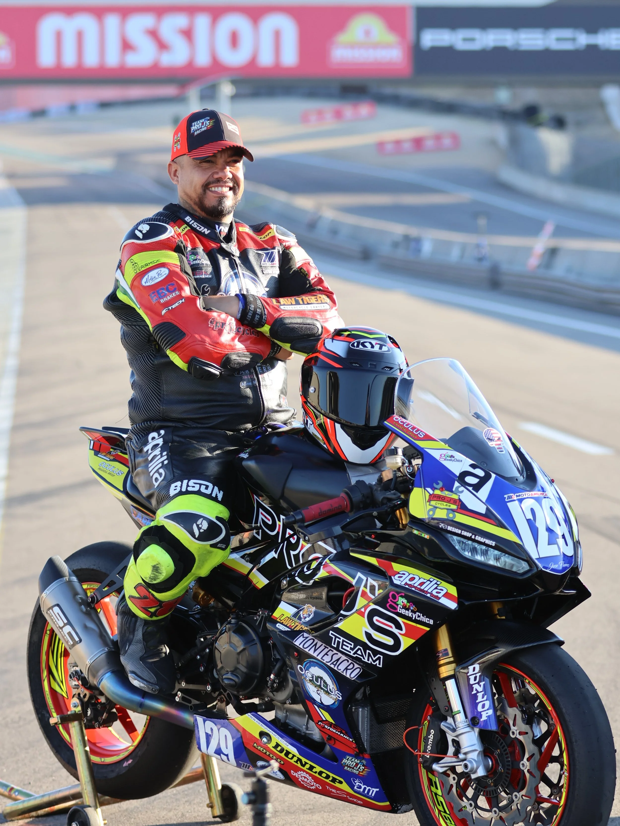 A man in a racing suit and helmet stands beside a race motorcycle on a race track, smiling.