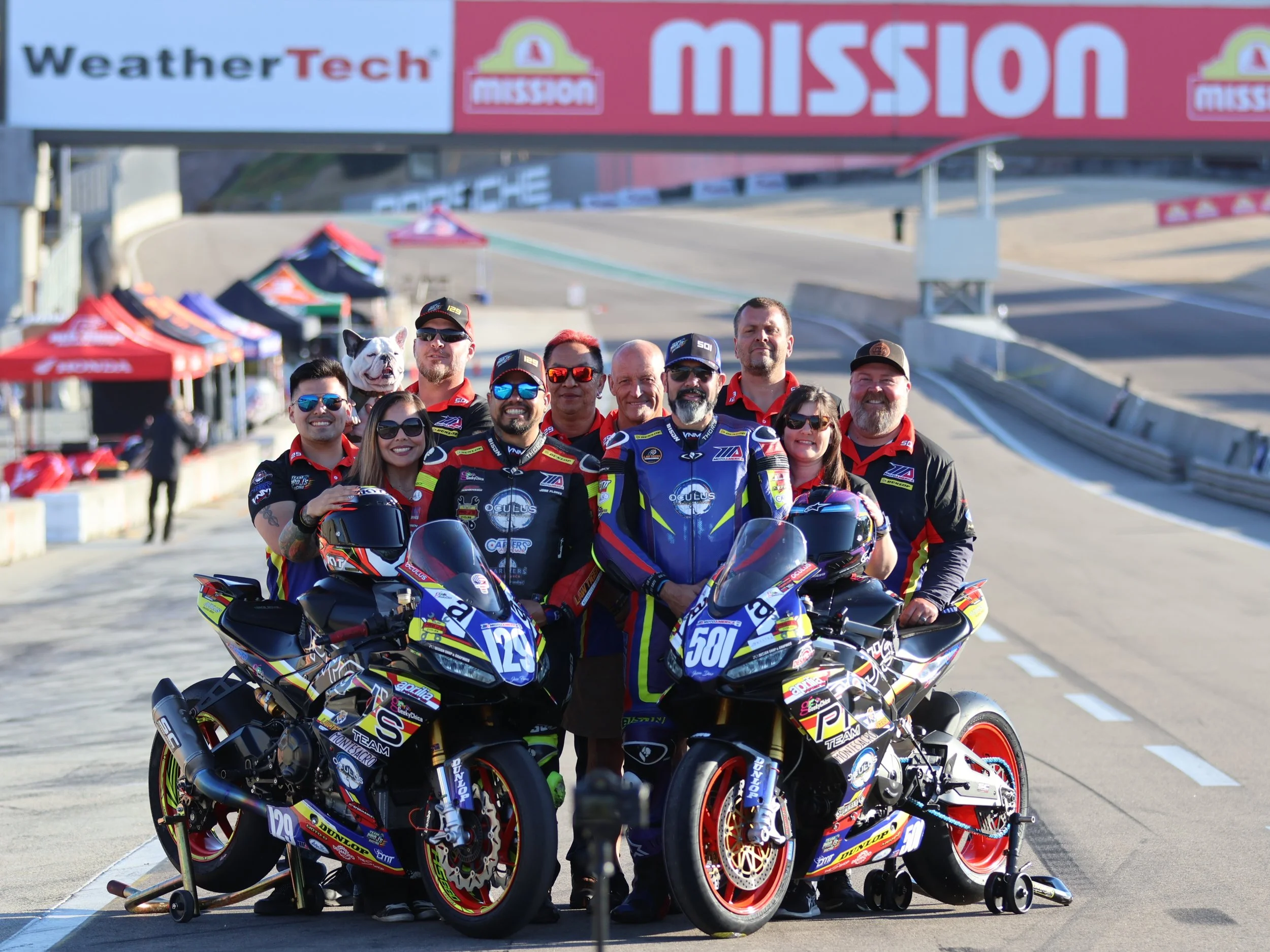 Motorsport team posing with racing bikes on a race track, under a banner that says 'MISSION', with pit tents and officials in the background.