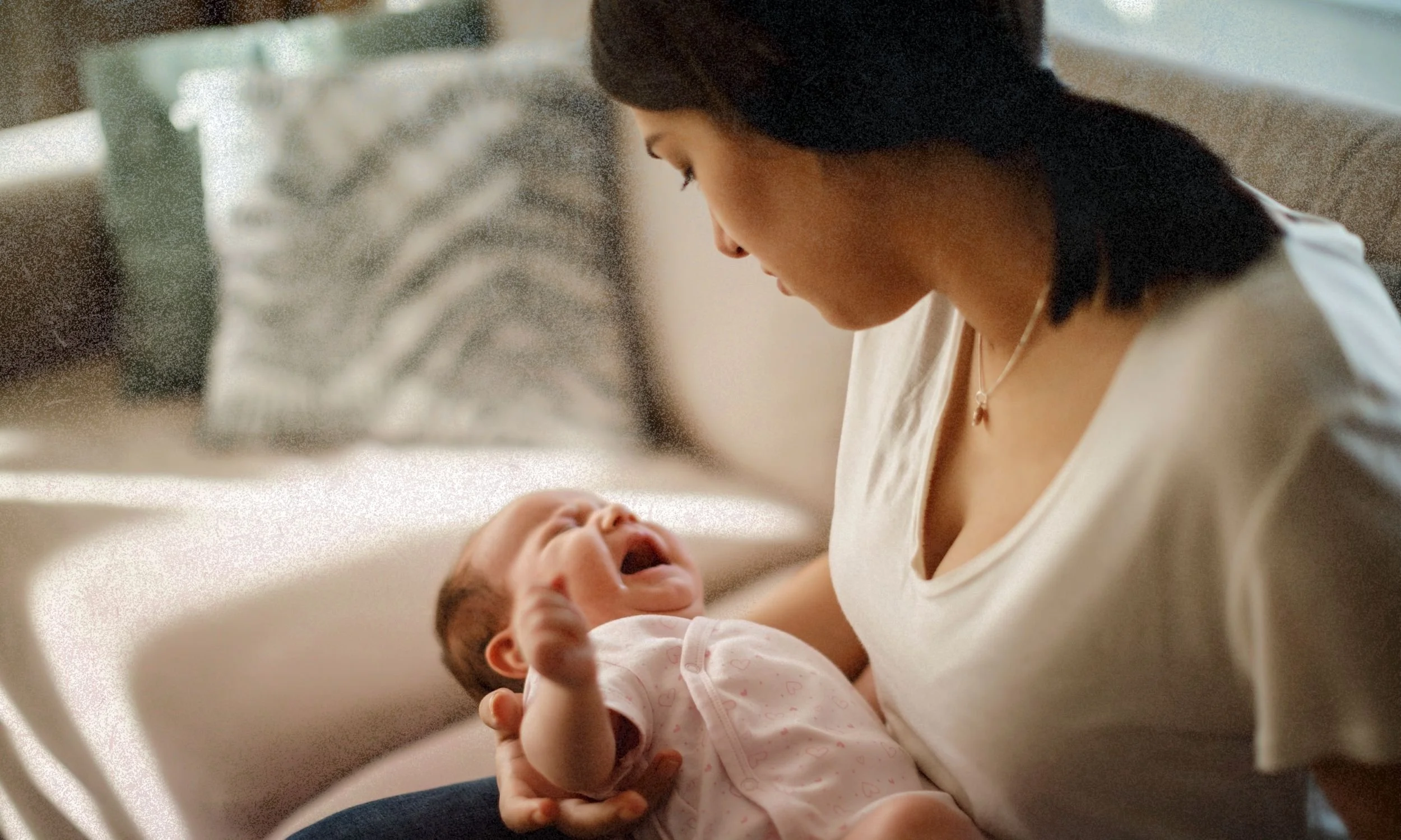 A parent in a white shirt holds a baby on a sofa, gently supporting the infant while looking down in a cozy, sunlit living room.