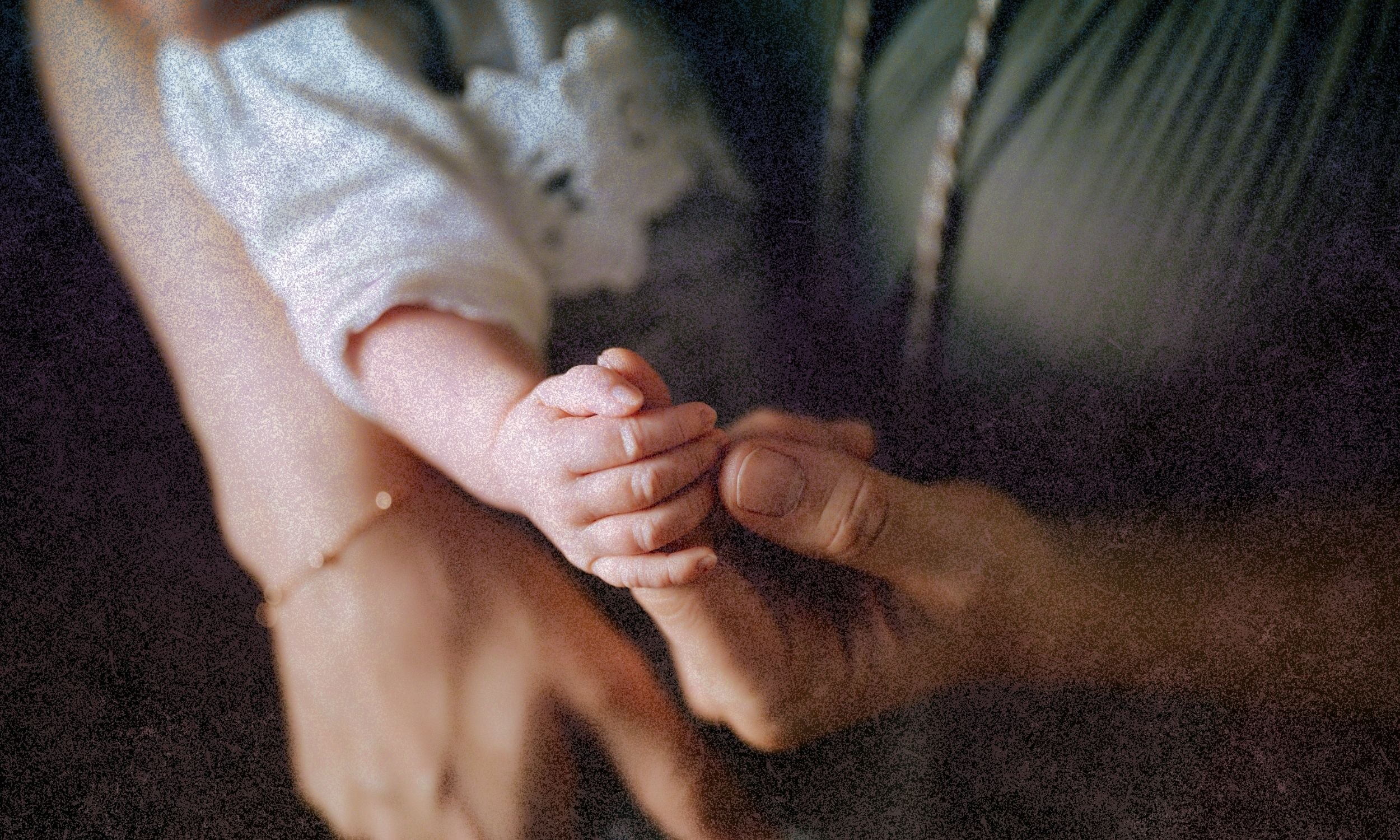 Close-up of an adult holding a child’s small hand while gently clasping wrists, with warm light and soft focus on a dark background.