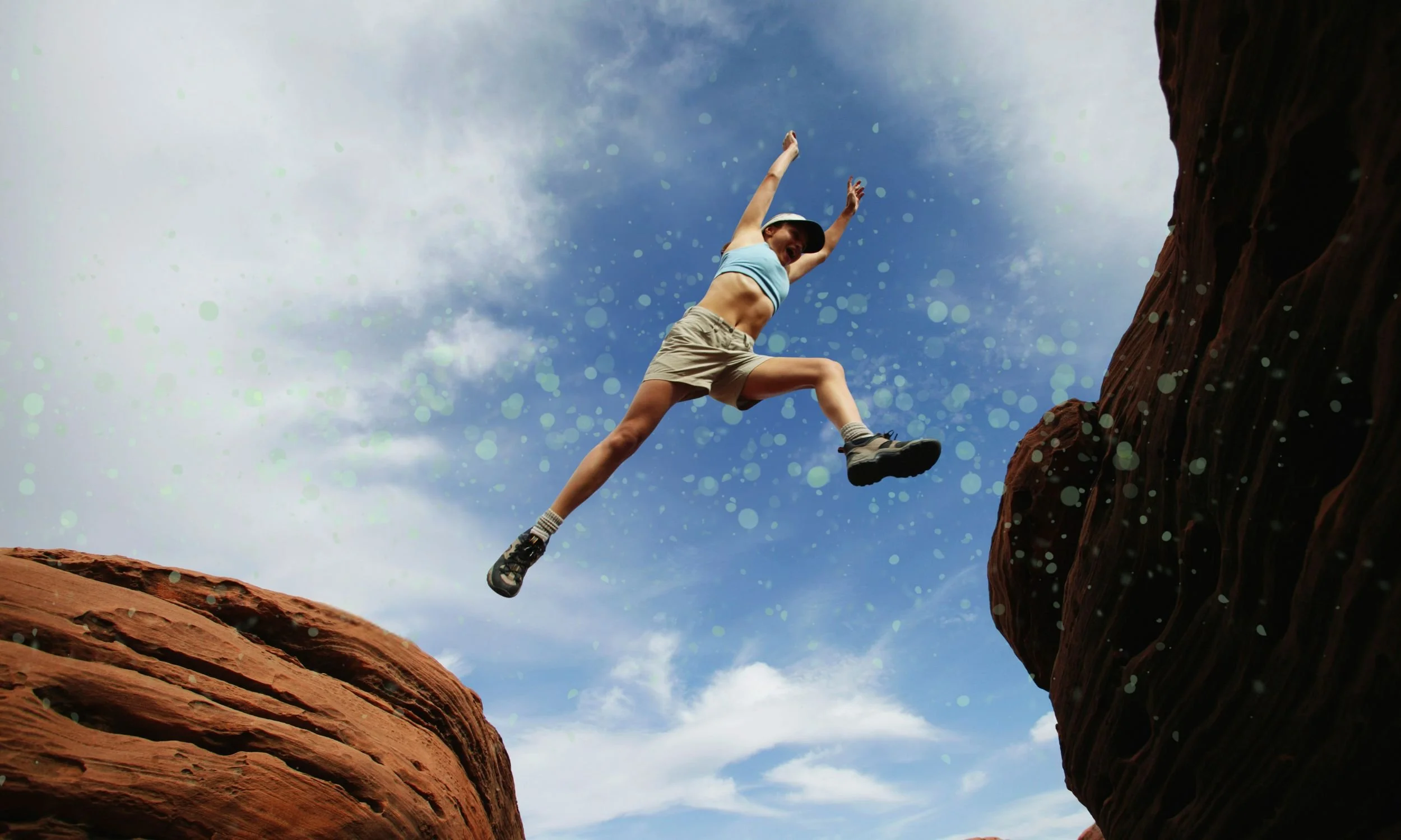 A person leaps across a gap between two large red rocks, with a blue sky and white clouds in the background.