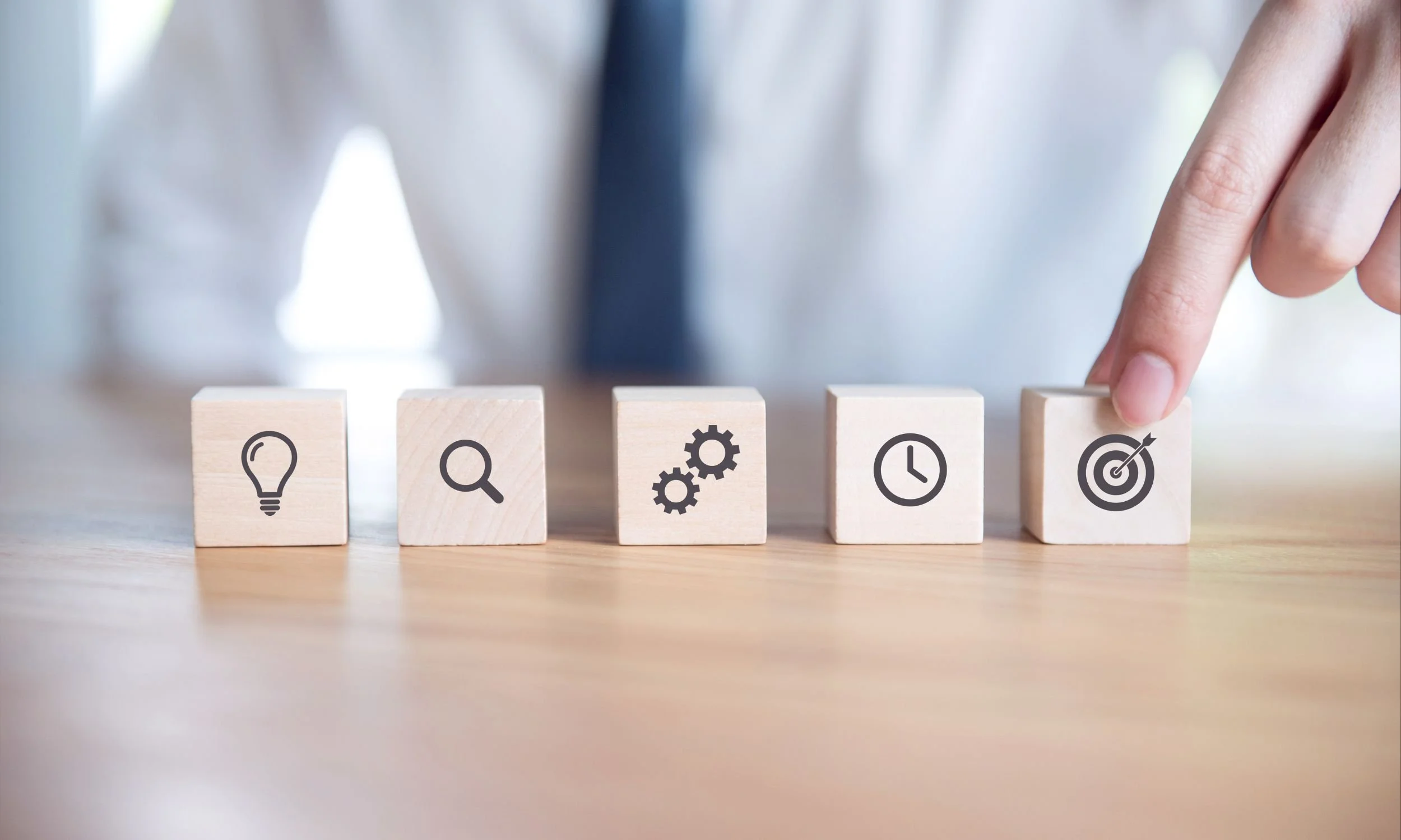 Person lining up 5 blocks of wood together. Each block has various logos printed on them: lightbulb, magnifying glass, gears, clock, and target.