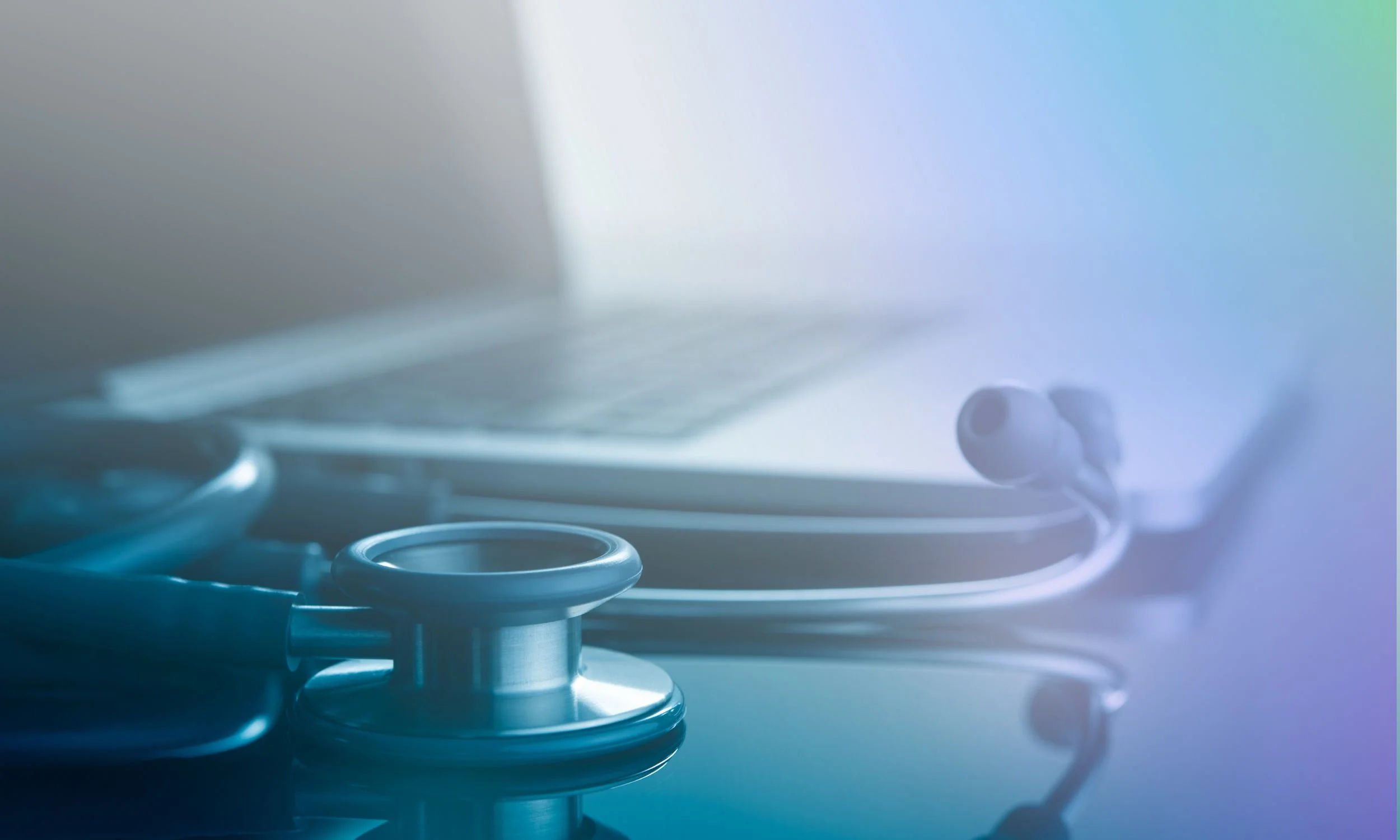 A stethoscope rests on a reflective surface in front of a blurred laptop, suggesting a connection between medical tools and technology.