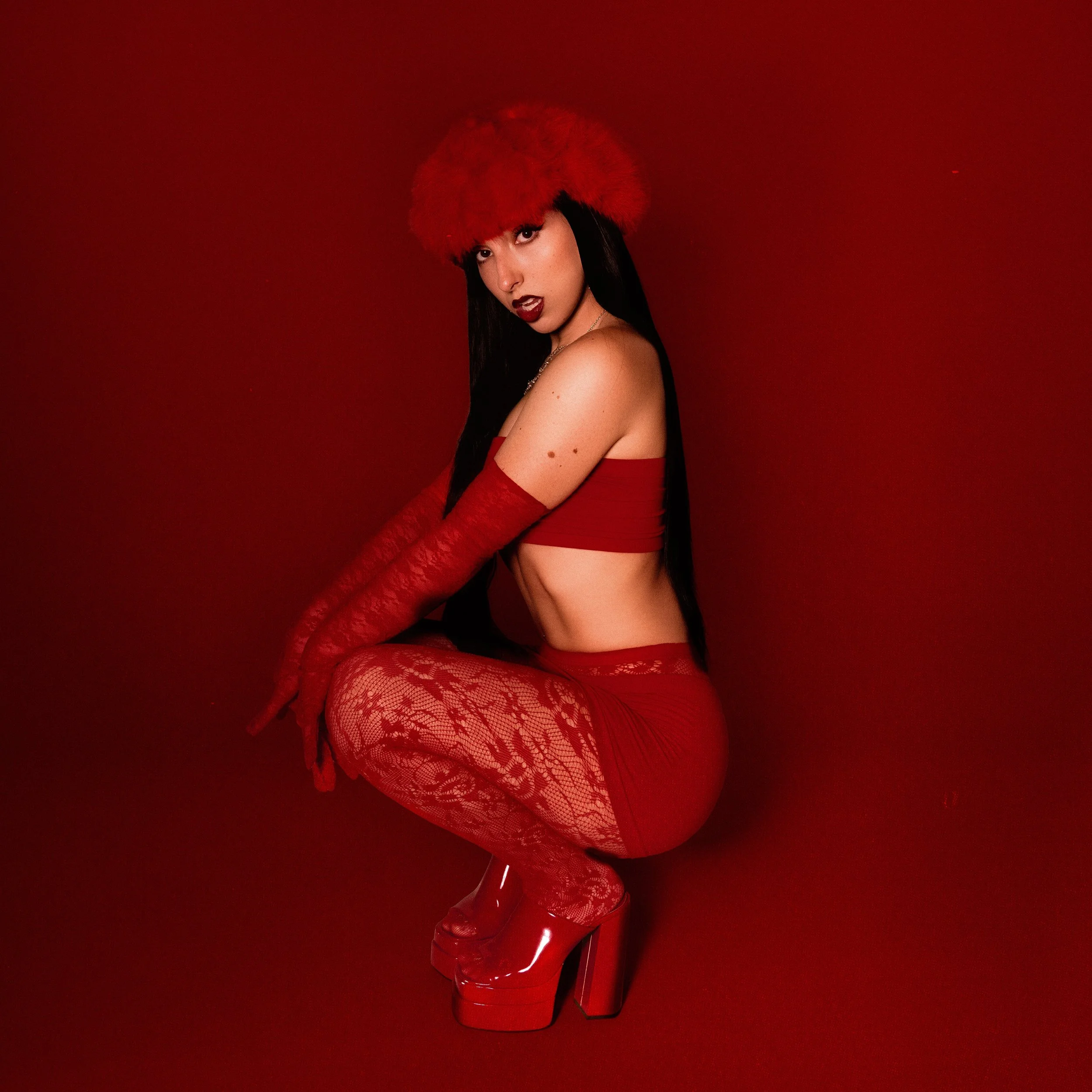 A woman dressed in red, wearing a furry red hat, lace gloves, lace stockings, high heels, and a red crop top, posing against a red background.