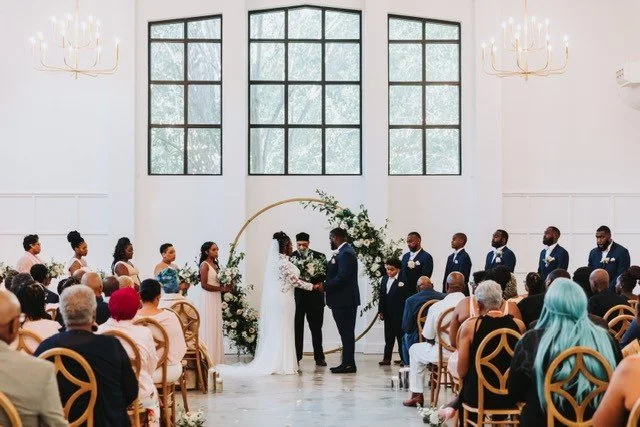 Indoor wedding ceremony with bride and groom exchanging vows, officiant present, wedding party standing beside, seated guests, large windows, floral arrangements, and chandeliers.
