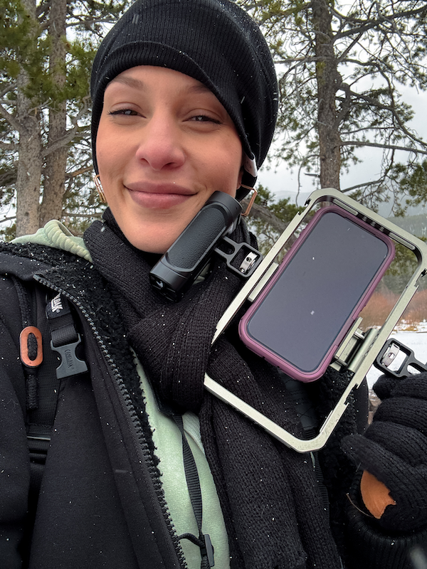 Rhi smiling while holding her phone cage up to her face as the snow falls down in RMNP.