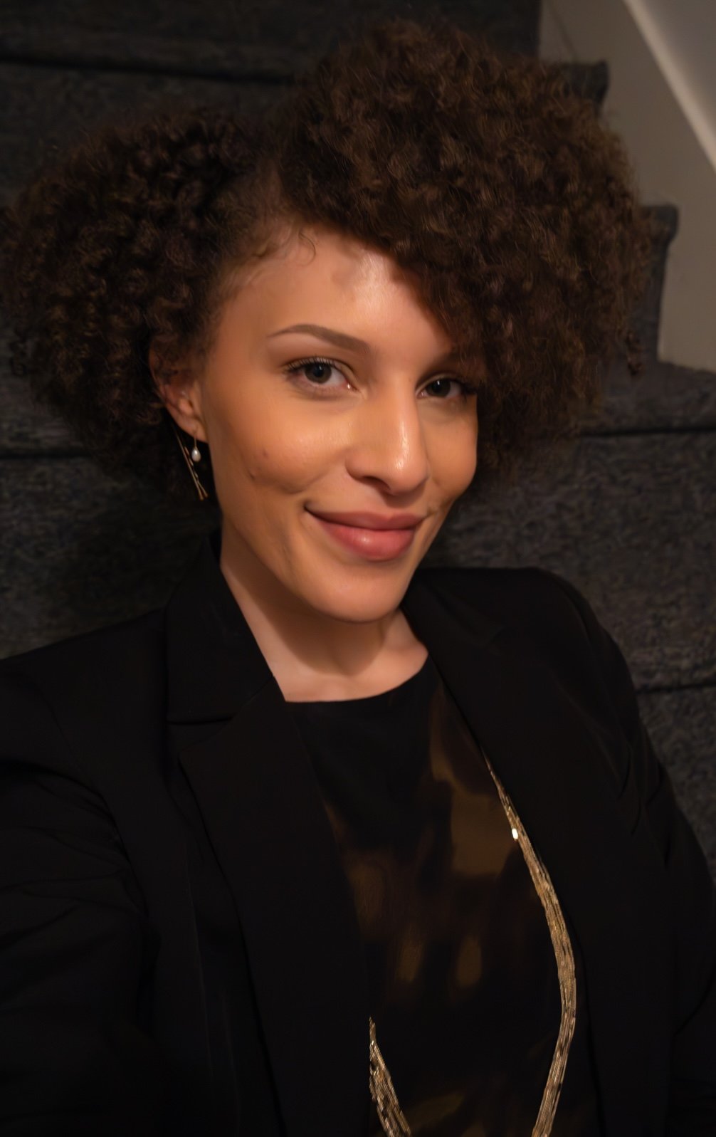 Rhi behind the scenes at a wedding she planned and coordinated in Niwot, CO. She is smiling at the camera with her fresh curly hair, black blazer, dainty pearl earrings, and black and green patterned dress.