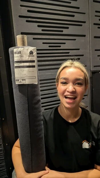 Young woman smiling and holding a large rolled-up gray wall panel in a home cinema room with acoustic tiles behind her.