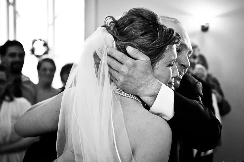 A bride and an elderly man, likely her father, hugging tightly during her wedding ceremony. The scene is tender and emotional, with guests blurred in the background.
