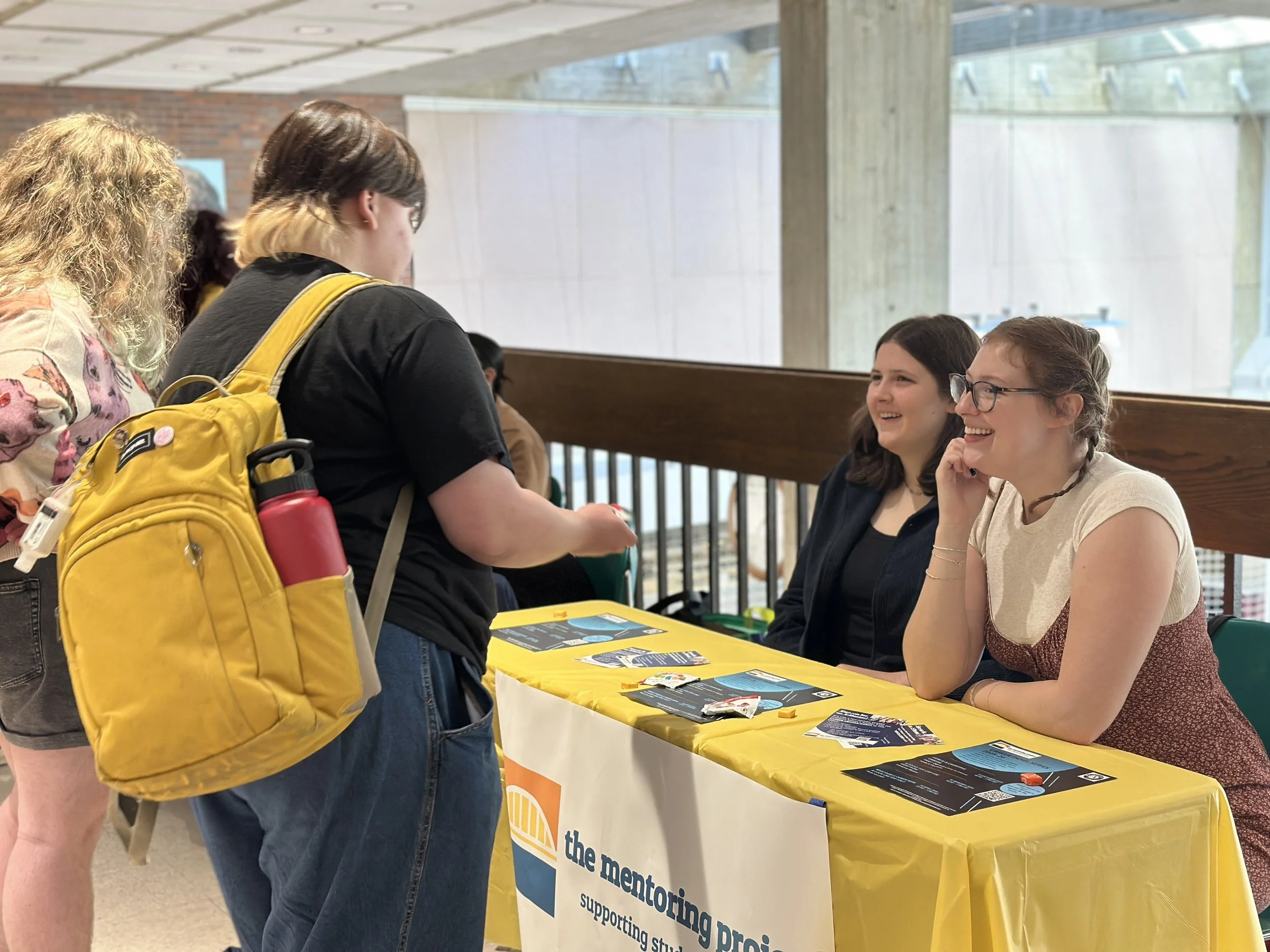Two women sitting behind a yellow table with flyers, smiling at a woman standing in front of them at an event, supporting mentoring students project.