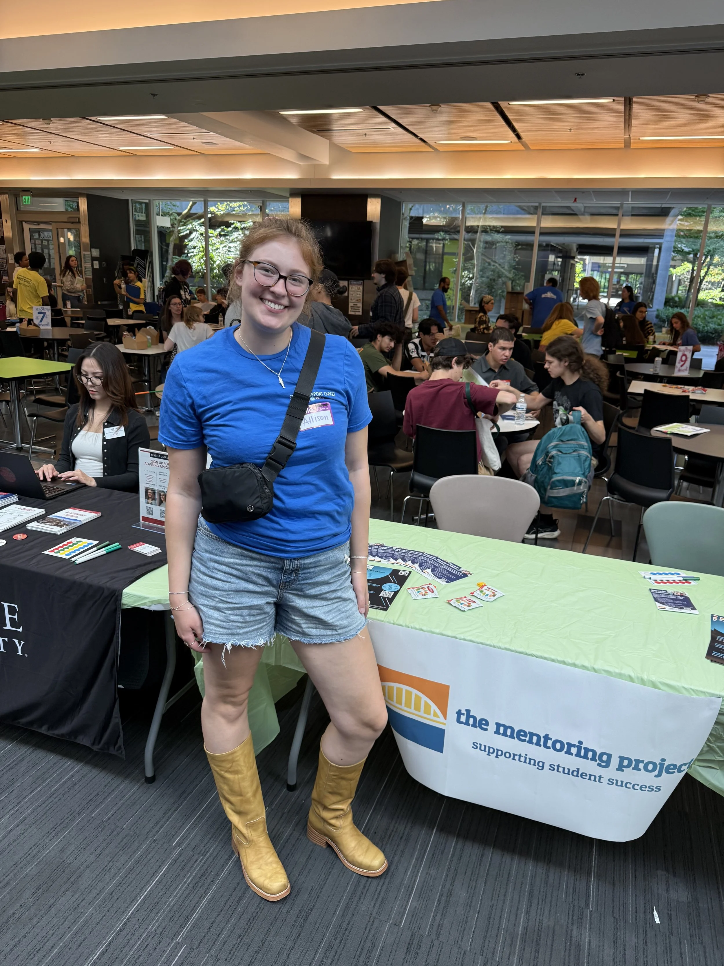 A woman with glasses, wearing a blue t-shirt, denim shorts, and yellow boots, standing at a table with a banner that reads 'the mentoring project supporting student success.' She is smiling in a busy indoor setting with many people in the background.