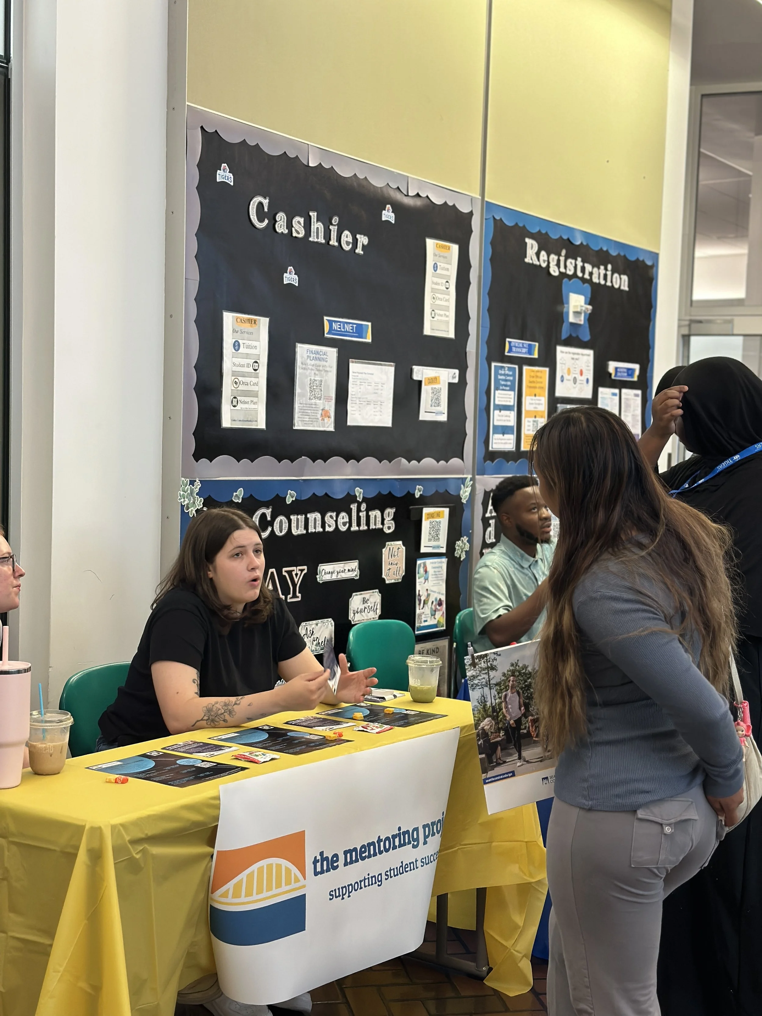 Students talking at a university mentoring program informational booth with black and yellow walls, displaying recruitment and counseling information.