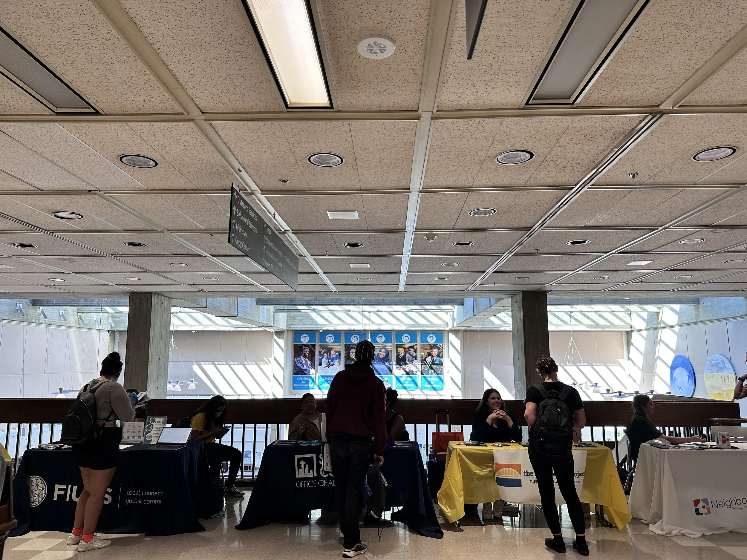 People standing and sitting at tables with banners in an airport terminal, with digital screens and architectural features in the background.