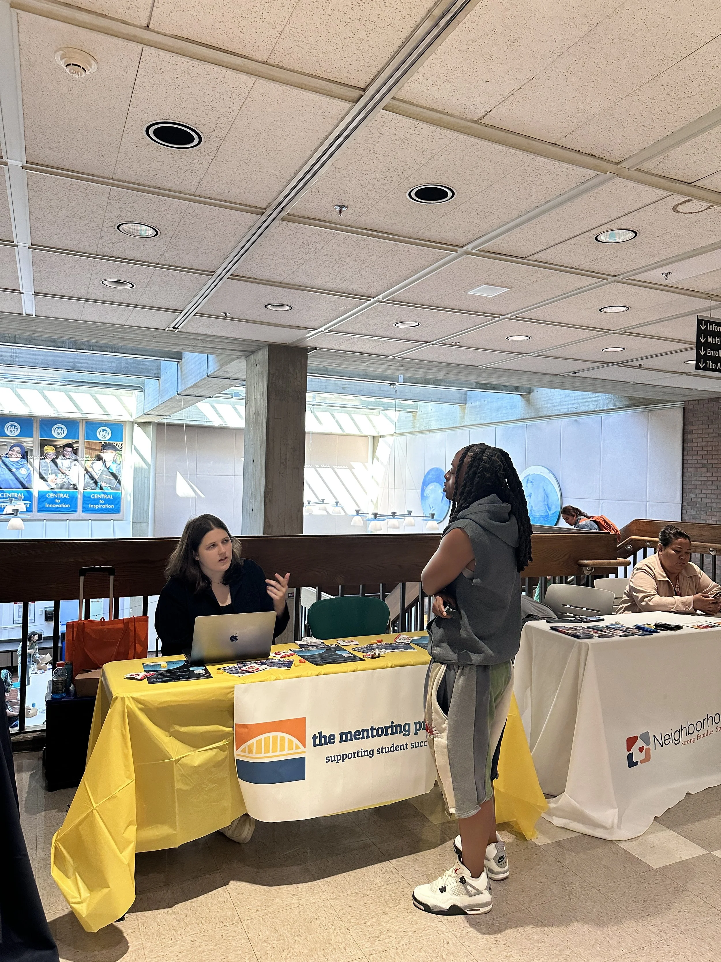 A woman in casual sportswear speaking with a woman sitting at a table with a laptop, at a career fair or informational event. The table has a yellow tablecloth with a banner that says 'the mentoring place supporting student success.' Other tables are