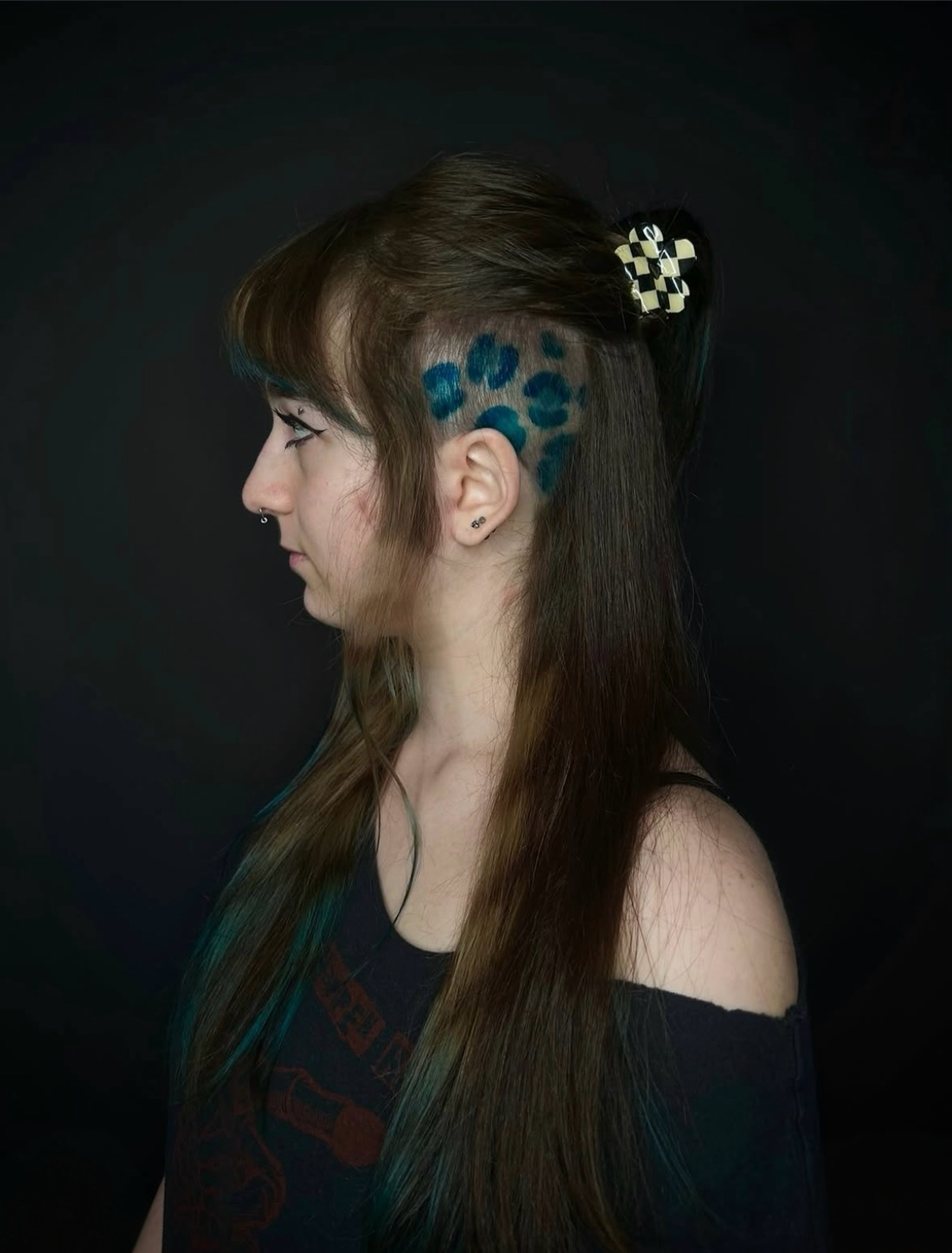 Side profile of a woman with partial shaved head, blue leopard print hair dye, long straight brown hair, black shirt, and checkered hair clip on dark background.