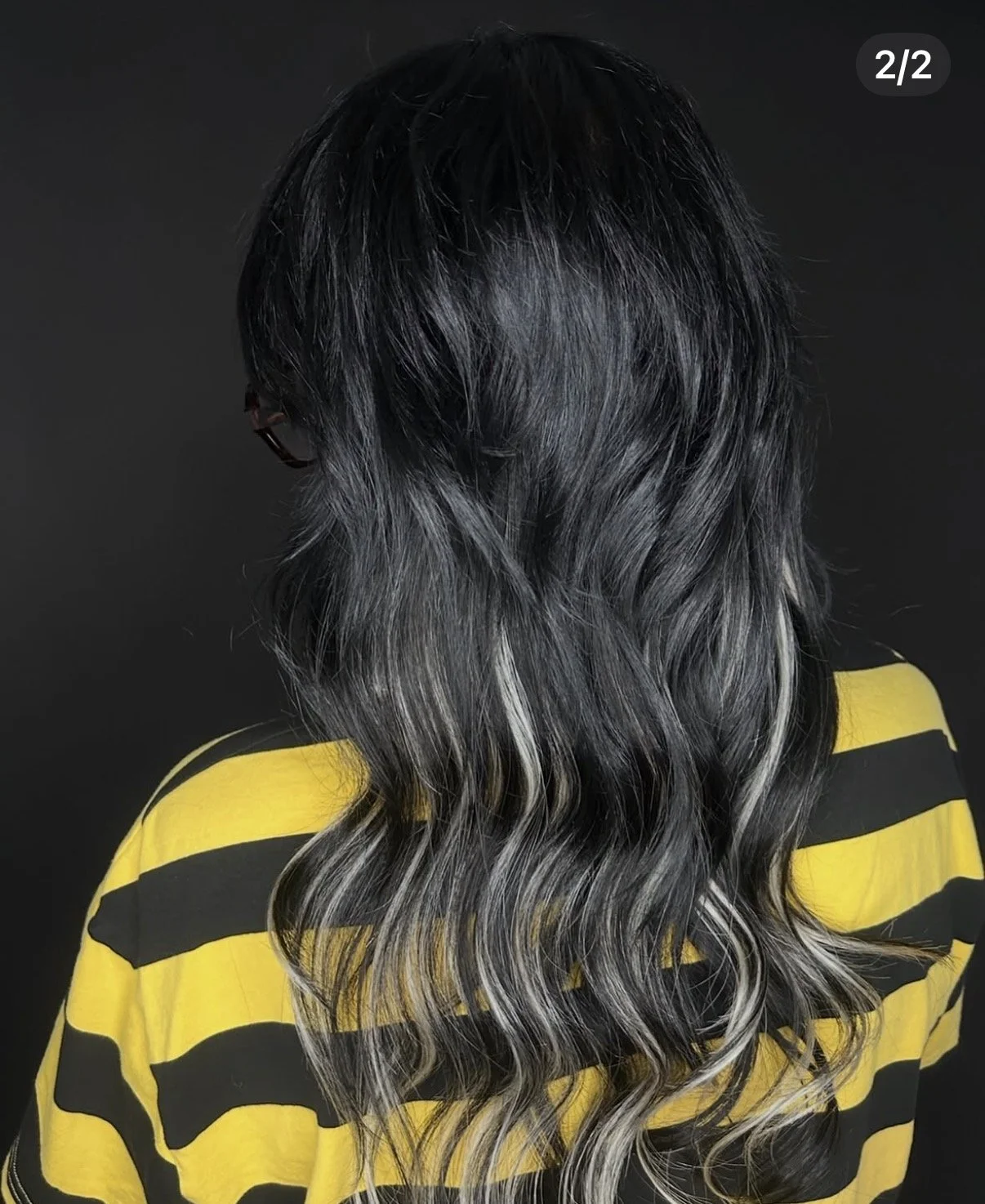 Back view of a person with long, wavy hair that is black at the roots and transitions to silver or gray at the ends, wearing a yellow and black striped shirt against a dark background.