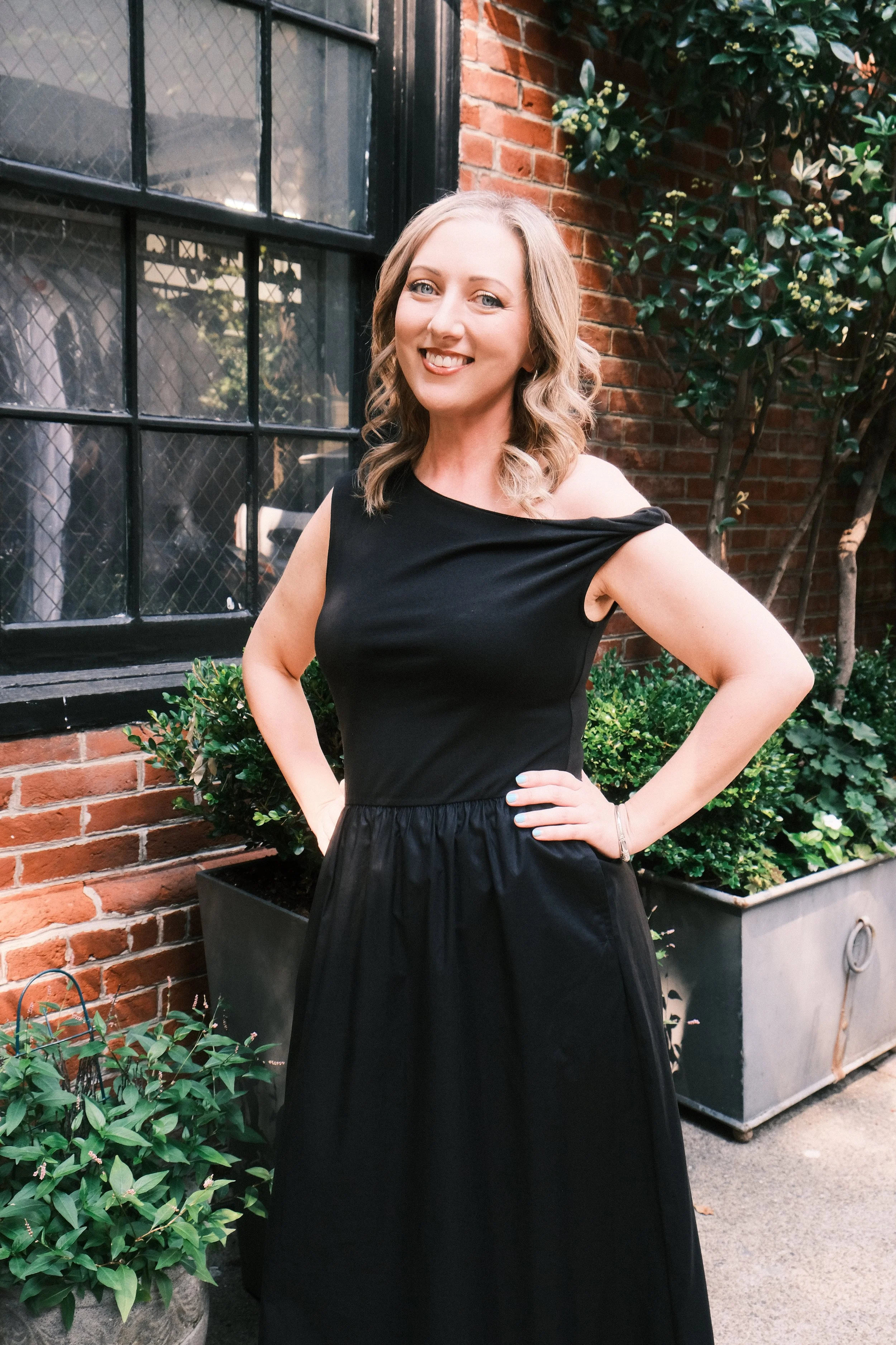 A woman wearing a black sleeveless dress stands outdoors near a brick wall with a large window. She has shoulder-length blonde hair and is smiling with her hand resting on her hip.