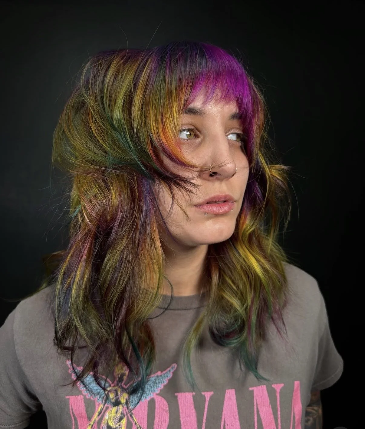 A woman with multi-colored hair including purple, pink, green, and orange streaks, wearing a gray T-shirt with a butterfly graphic and pink lettering. She has a nose ring and a small lip piercing, and is looking slightly to the side against a black b