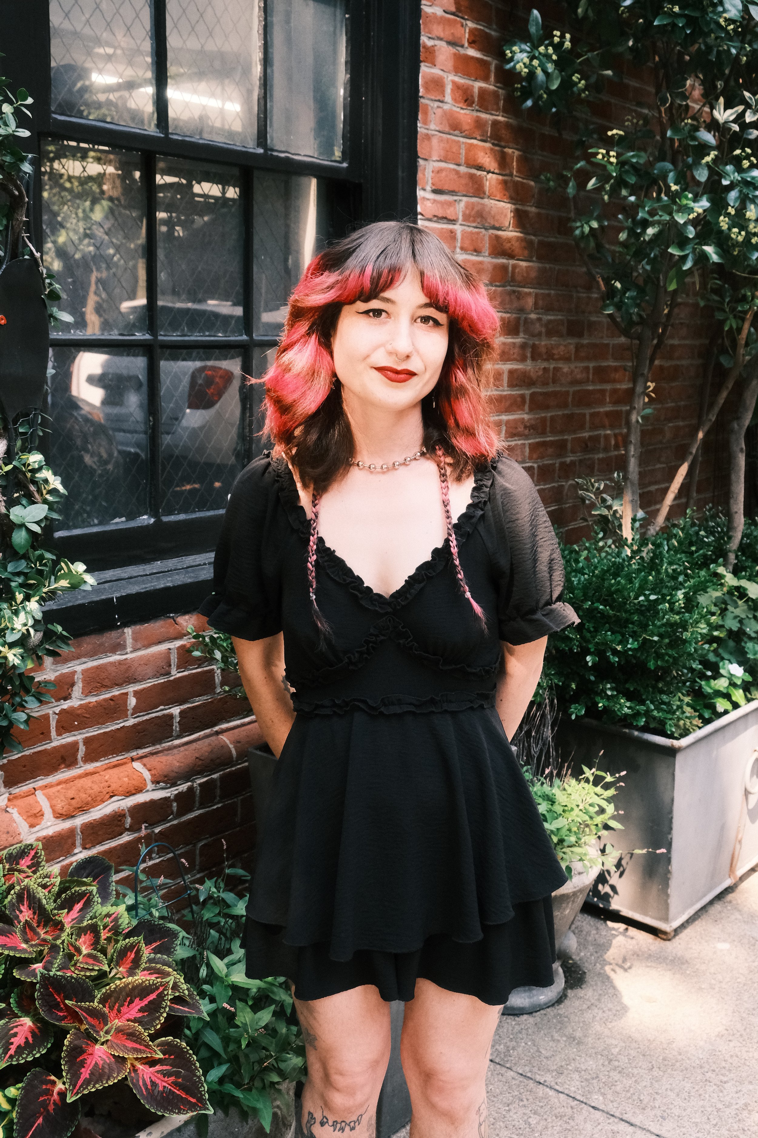 A woman with wavy, shoulder-length hair dyed with pink tips, wearing a black dress with ruffled details, standing outdoors near a brick wall and leafy plants, with a window behind her.