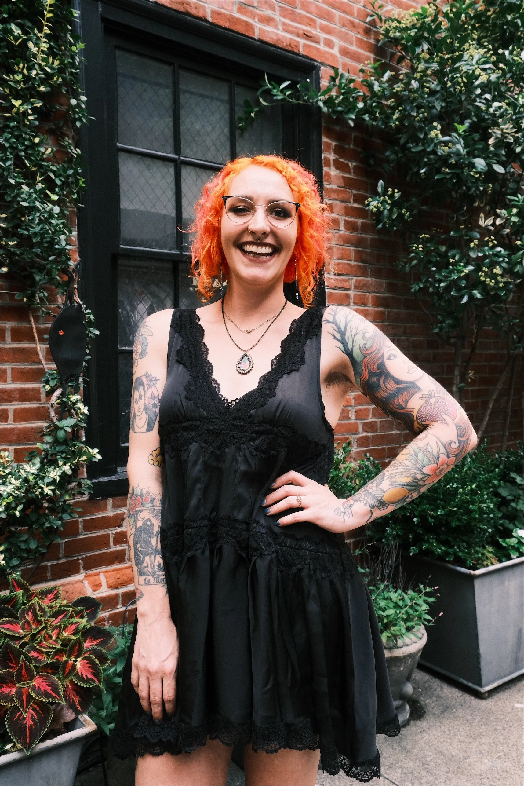 A woman with curly orange hair and tattoos on her arms, smiling and wearing glasses, a black lace-trimmed dress, and jewelry, standing outdoors in front of a brick building with green plants.