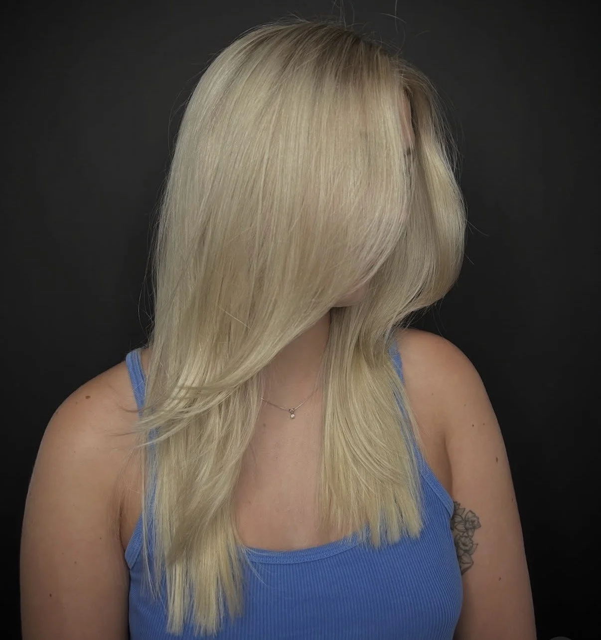 A woman with blonde hair and a blue tank top, her face is obscured by her hair, against a black background.