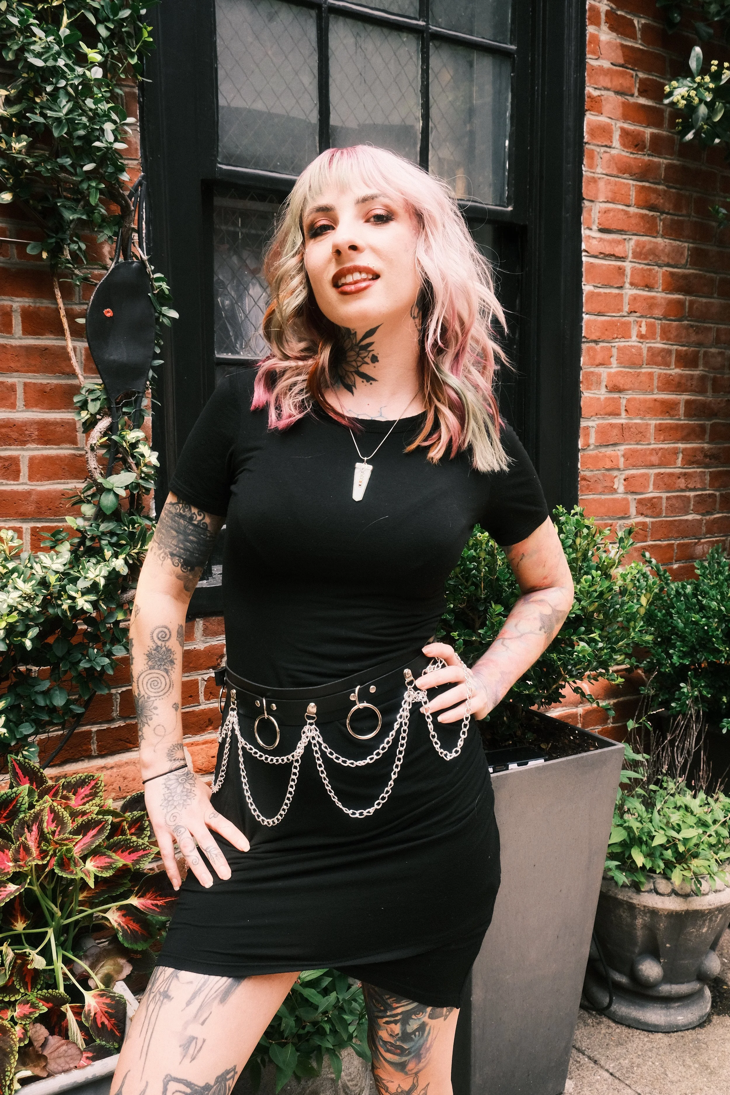 A woman with pink and blonde hair, tattoos, and dark makeup standing outdoors in front of a brick building and plants, wearing a black dress and chain accessories.