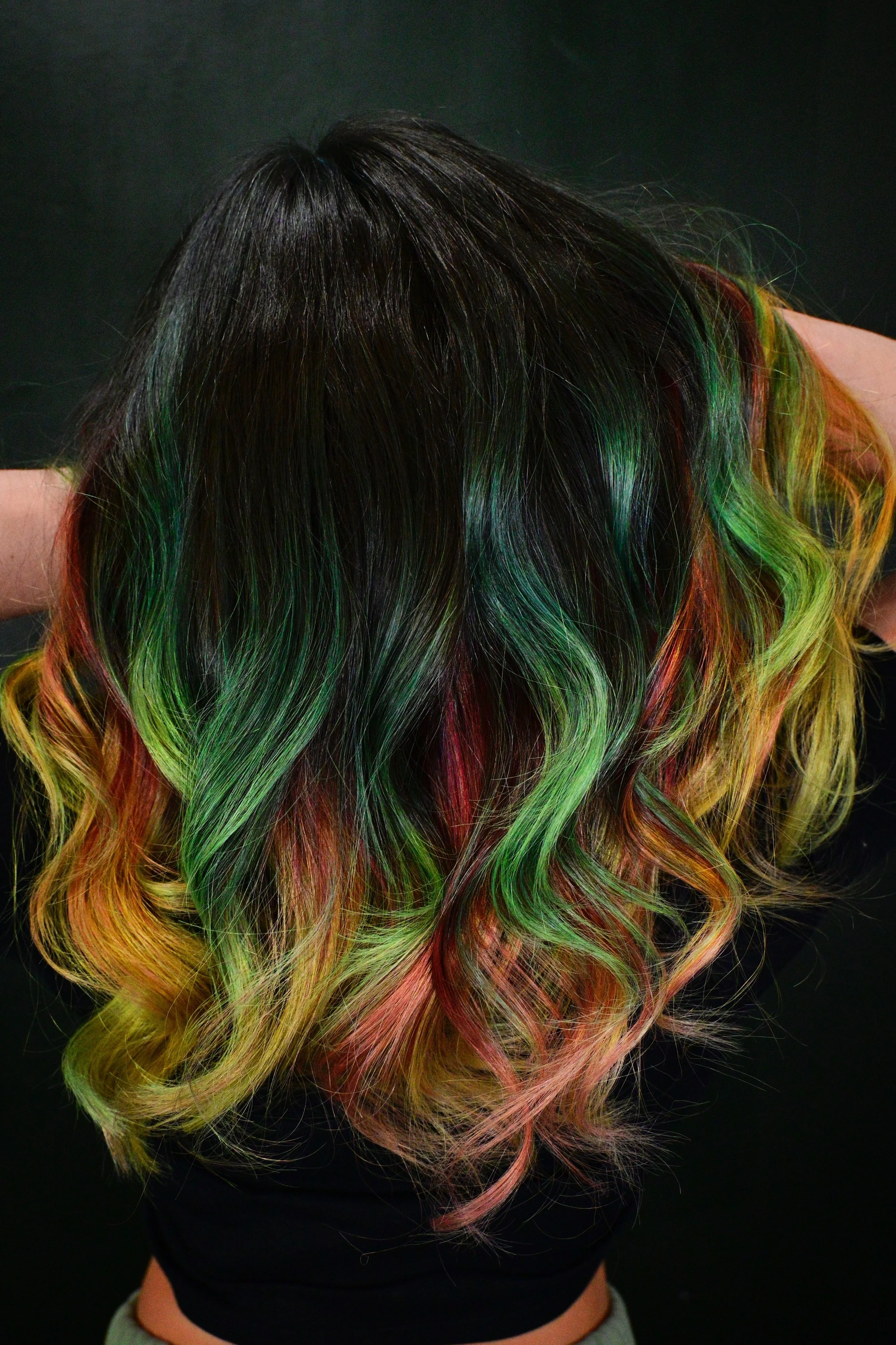 A person with long, wavy hair dyed in rainbow colors, including green, yellow, orange, and red, against a dark background.