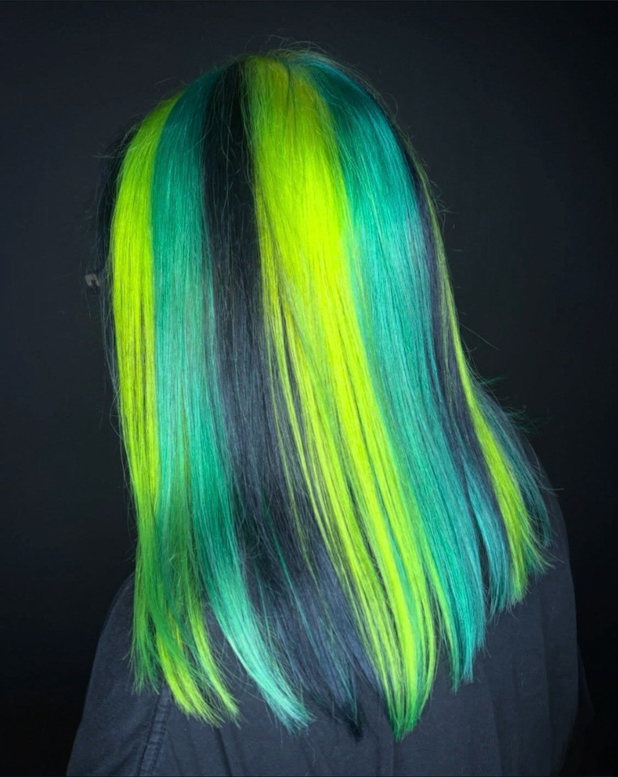 Person with long, straight hair dyed in neon green, yellow, and blue colors against a black background.