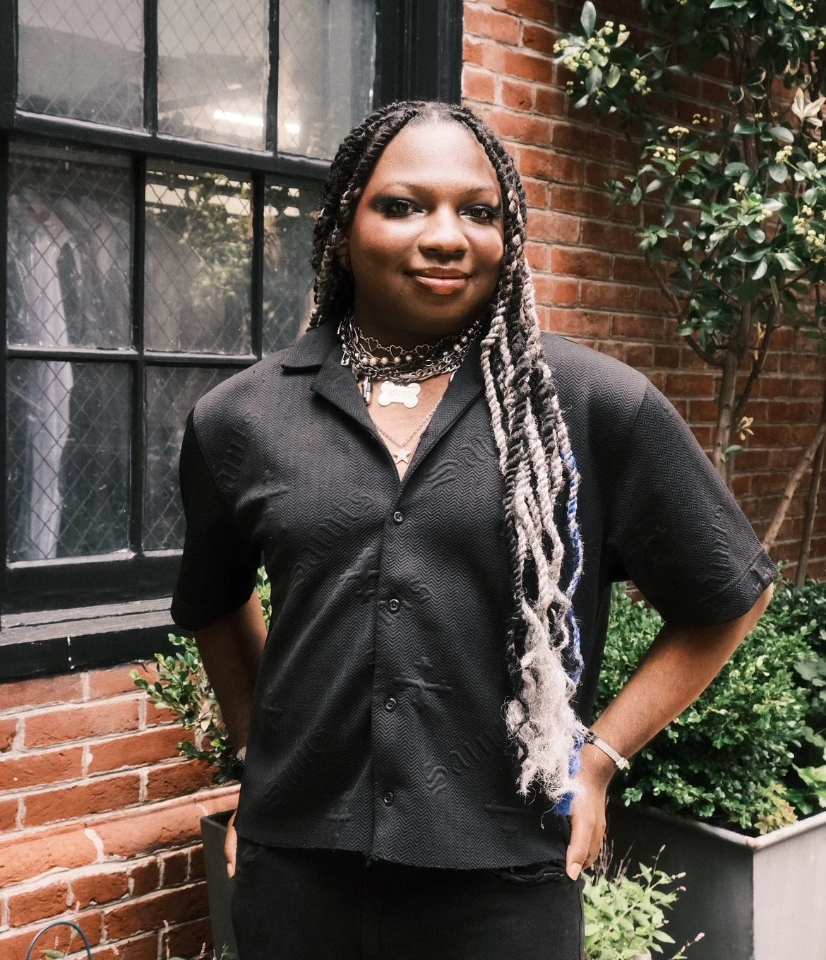 A woman with long braids standing outdoors in front of a brick wall and large window. She is wearing a black short sleeve button-up shirt and layered necklaces.