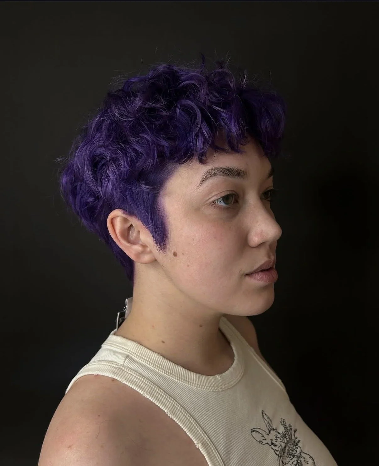 A young woman with short, curly purple hair wearing a light-colored sleeveless top with a deer illustration on it, against a dark background.