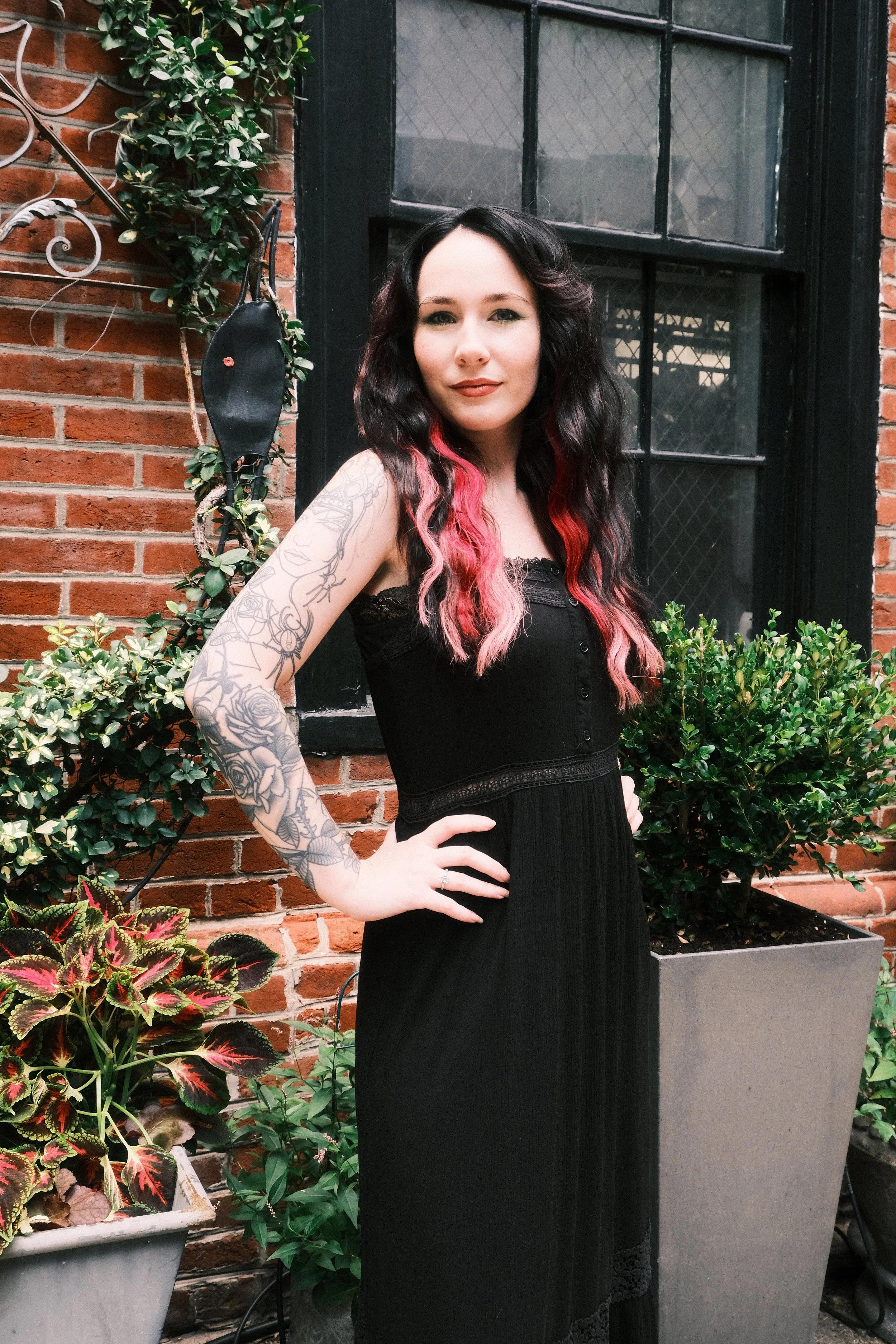 A woman with tattoos on her left arm, dark wavy hair with pink tips, wearing a black sleeveless dress, standing outdoors in front of a brick wall with large window and potted plants.