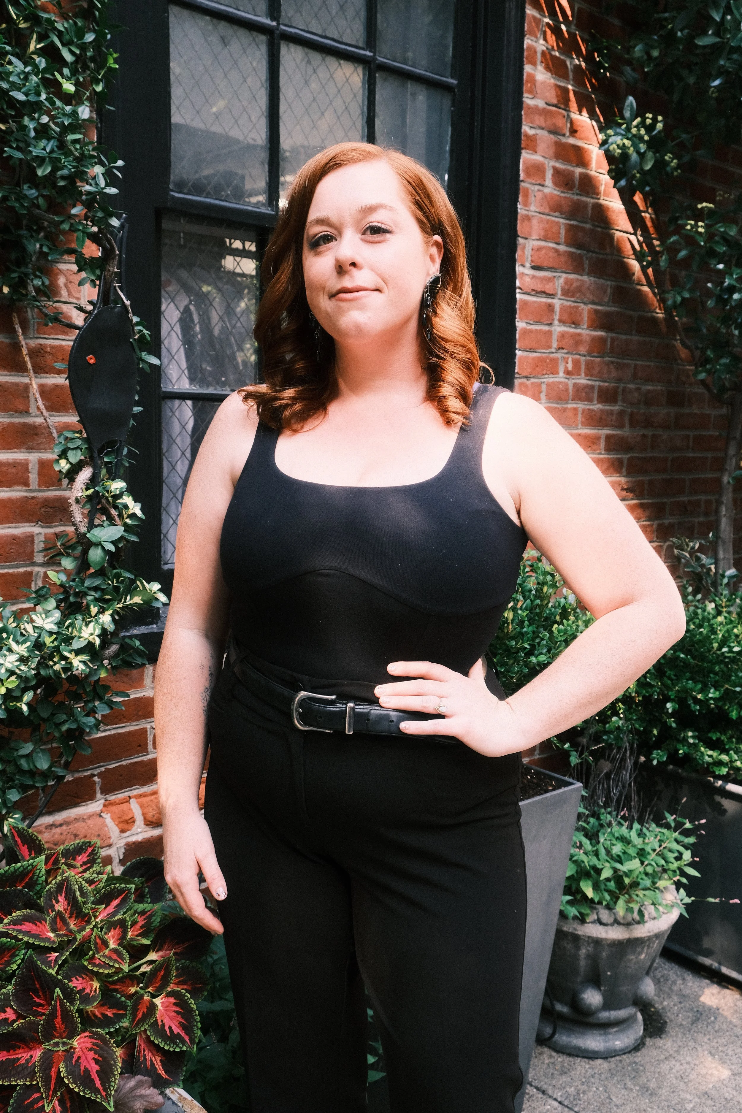 A woman with red hair wearing a black sleeveless top and black pants, standing outdoors against a brick wall and surrounded by potted plants.