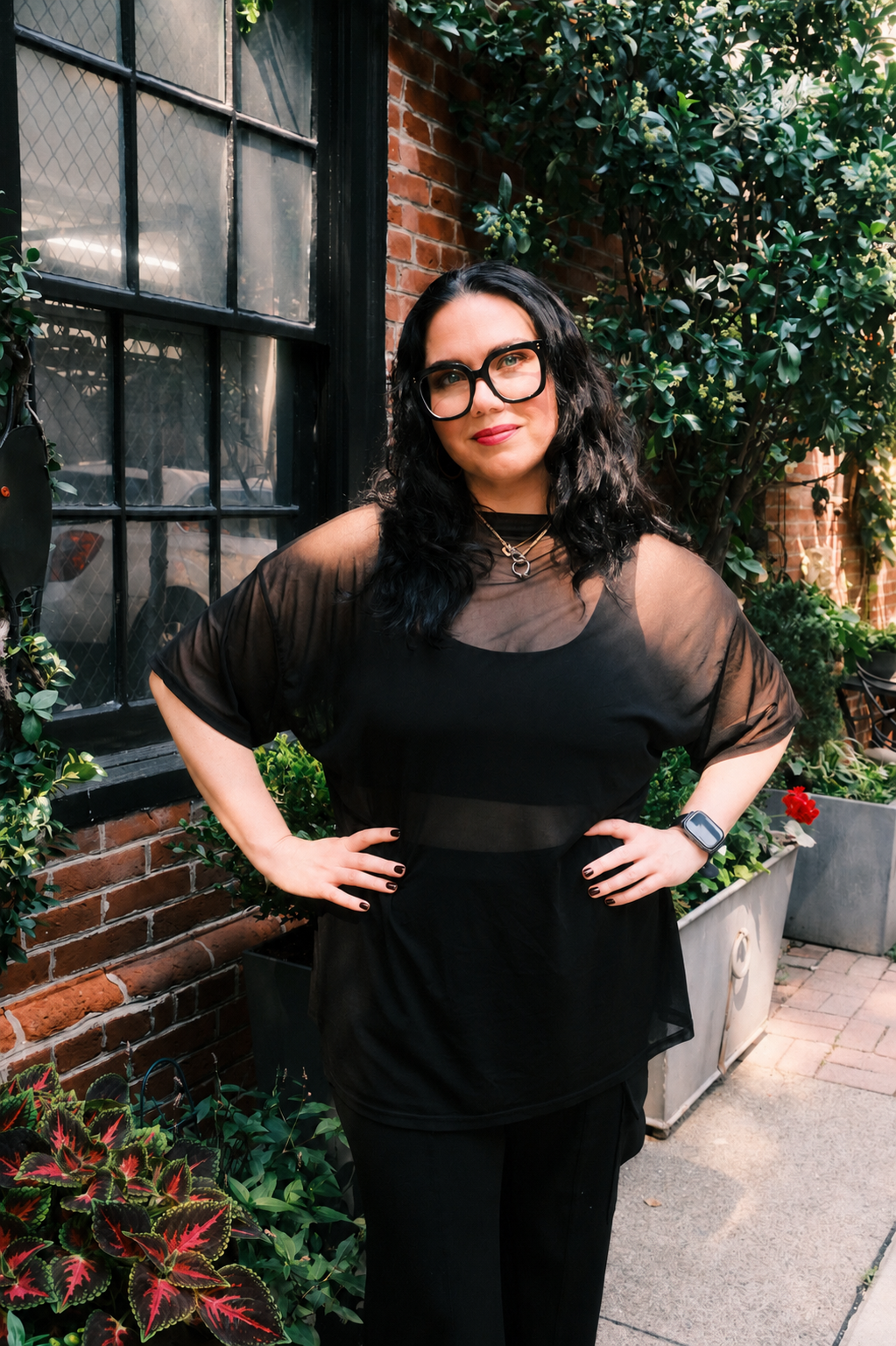 A woman with black curly hair, glasses, and a smartwatch standing outdoors next to a brick wall and lush greenery. She is wearing a black dress with sheer sleeves, a silver necklace, and has her hands on her hips.