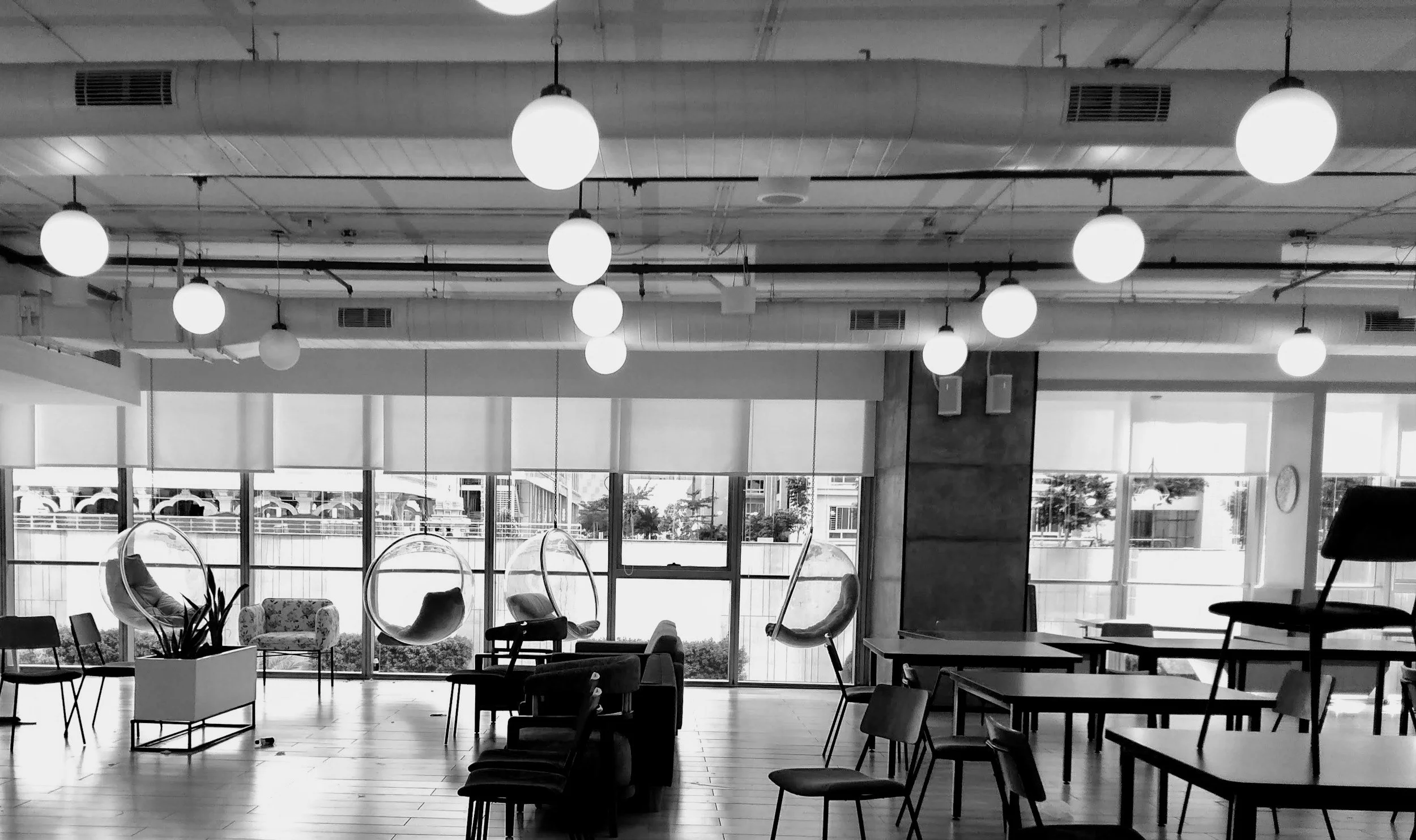 A modern lounge area with large windows, hanging globe lights, and a variety of chairs and tables, including hanging bubble chairs and potted plants.