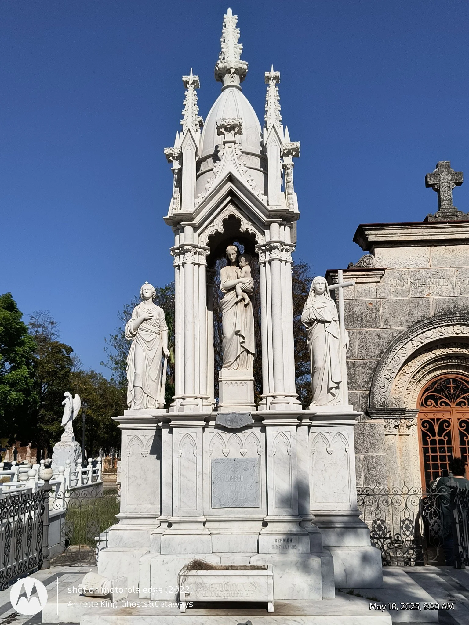 White marble monument with religious sculptures, including a central figure holding a child, flanked by two women. Ornate spires and cross atop. Part of a church or cemetery, with fencing and trees in background.