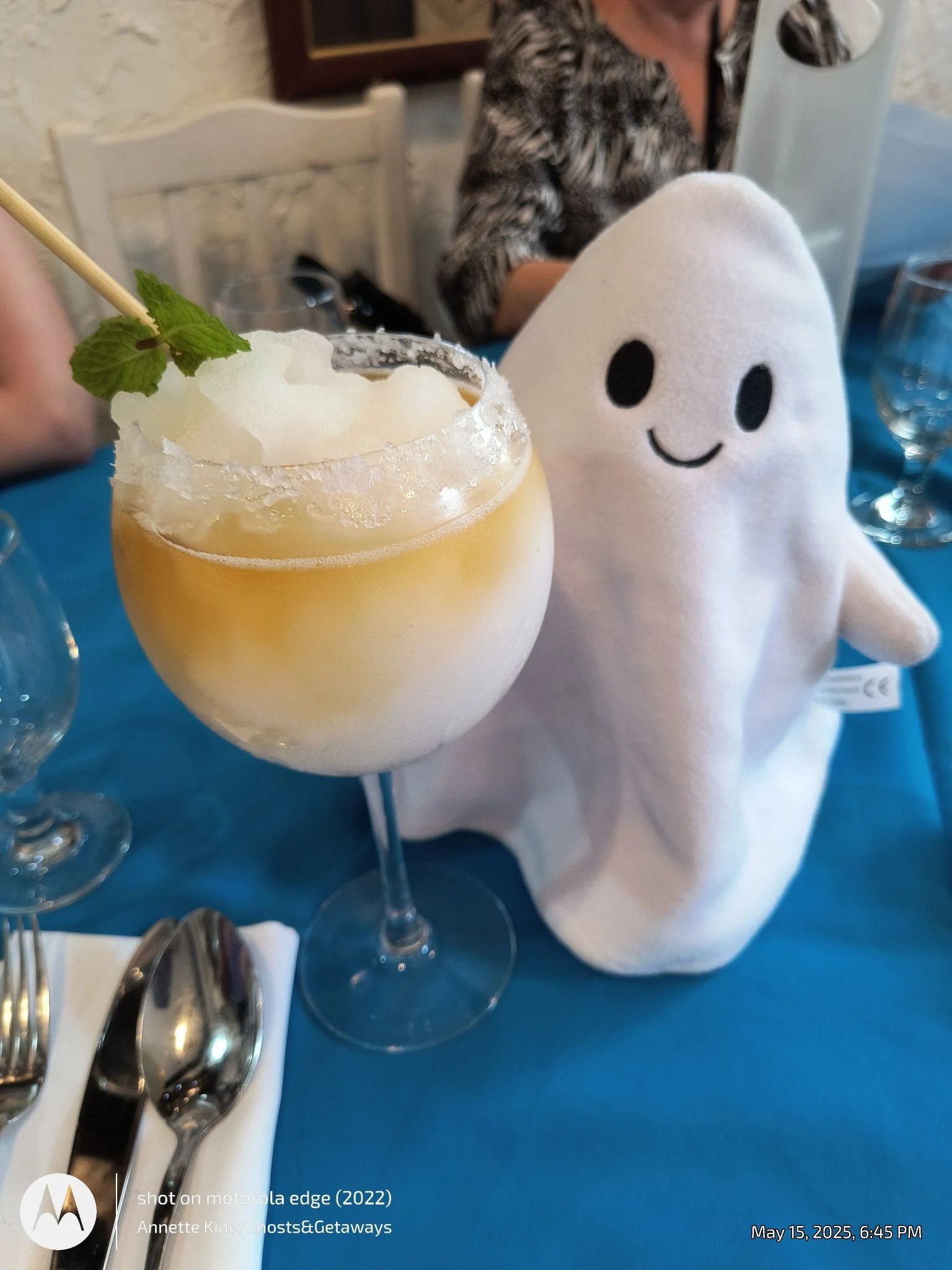 A close-up of a wine glass filled with a yellow beverage, topped with a ginger root garnish and a mint leaf, with a stuffed toy character with a smiling face, white body, and black eyes and mouth on a table with a bright blue tablecloth, and a woman 