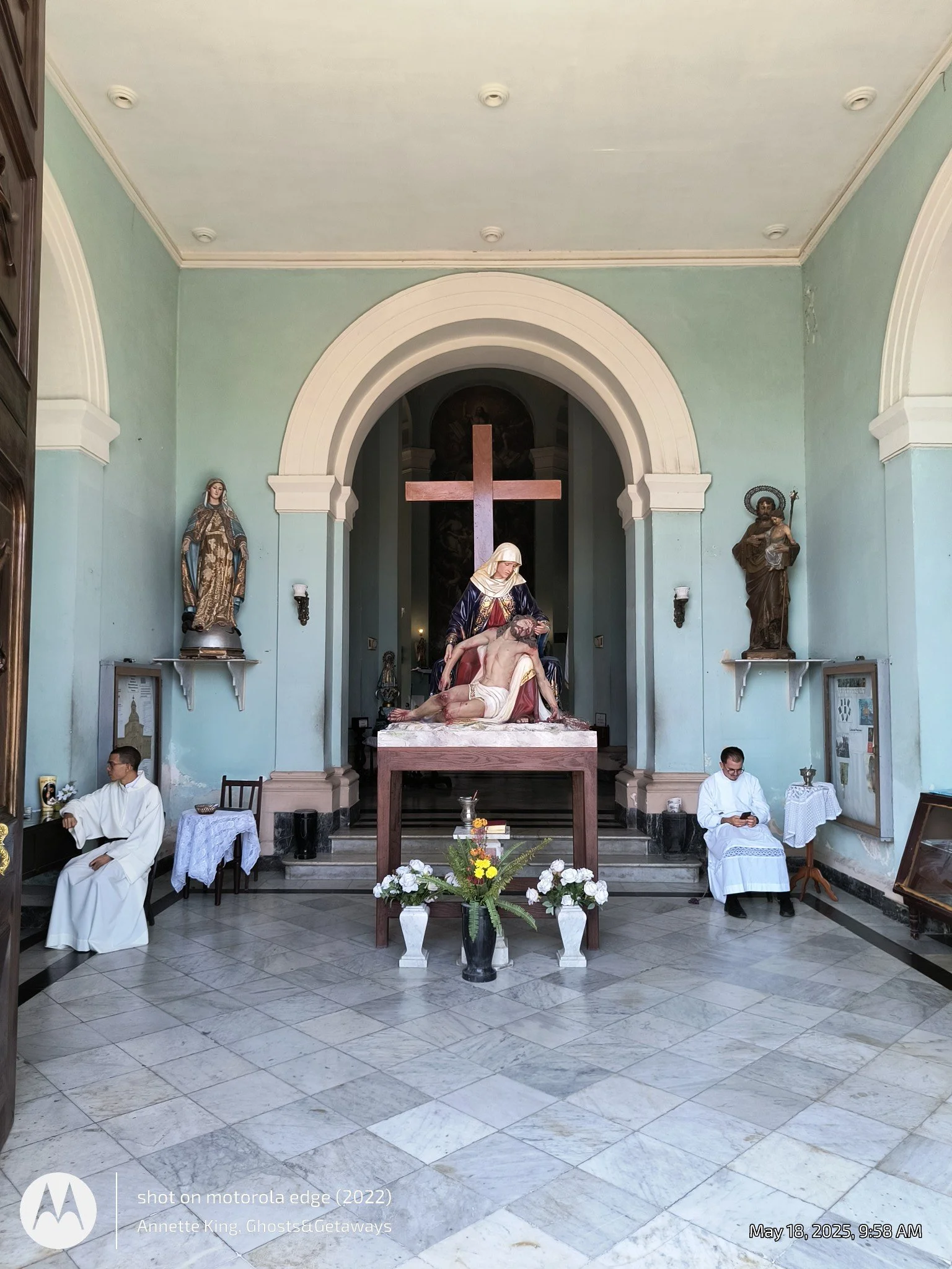 Interior of a church with a religious statue and altar, two men in white robes seated, one on each side, and a large wooden crucifix hanging above the altar.