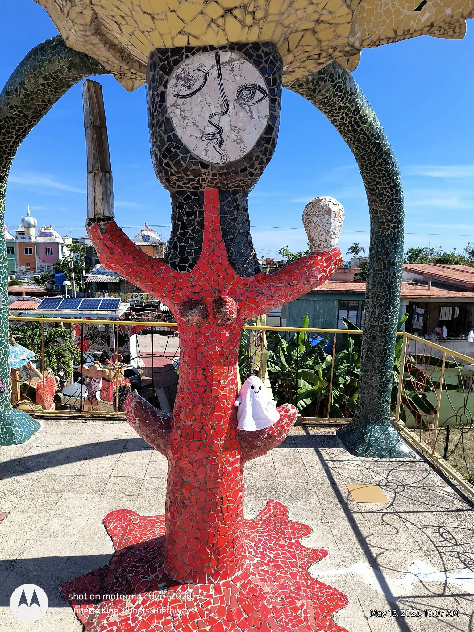Colorful mosaic sculpture of a woman with an abstract face, red dress, sitting cross-legged, holding a mirror, with another similar statue in the background, set against a cityscape with rooftops and a clear blue sky.