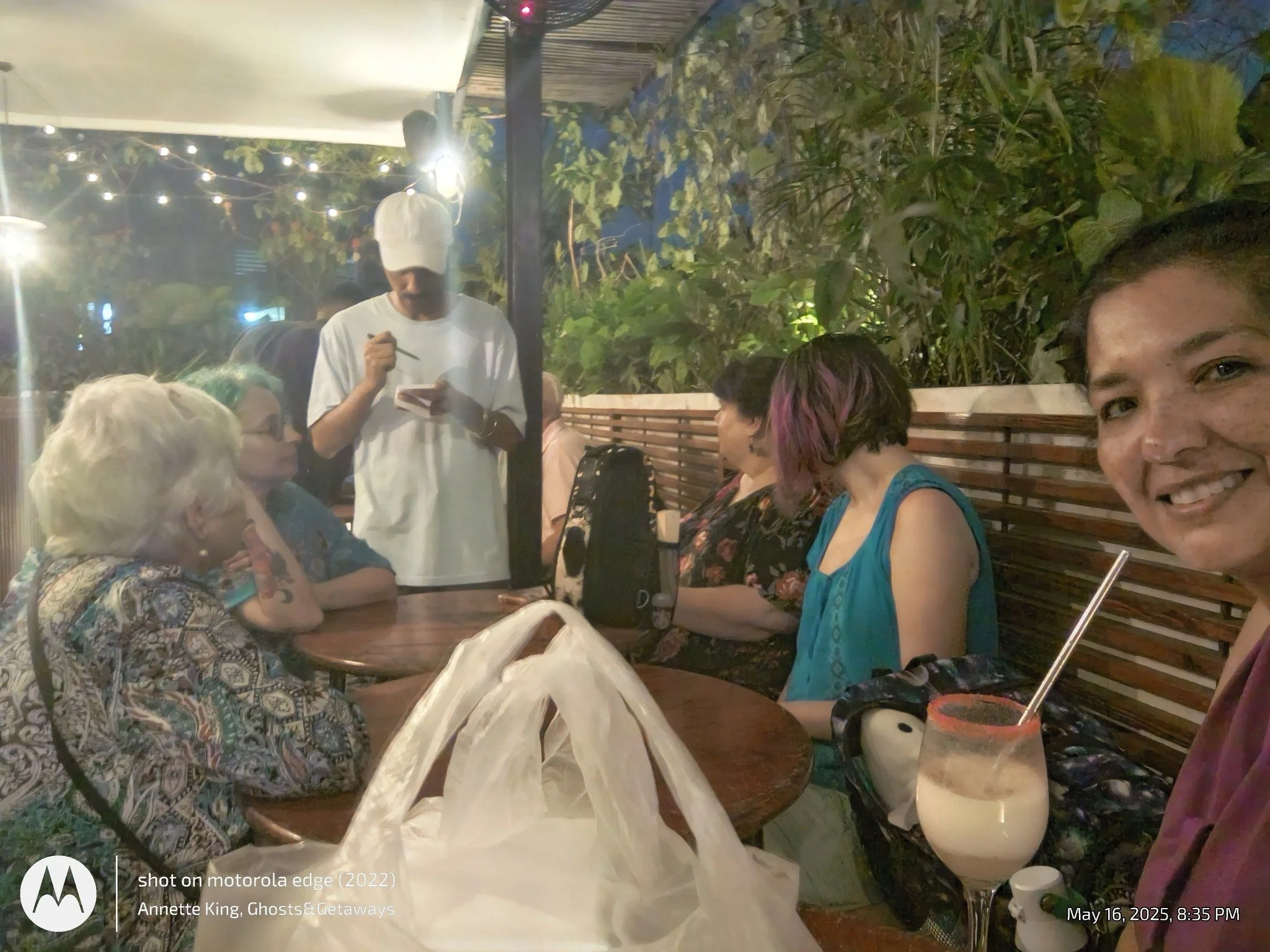A group of people sitting around a table at an outdoor gathering during the evening. There are string lights and greenery in the background, with a man wearing a white baseball cap and white shirt standing and writing on a notepad. There are visible 