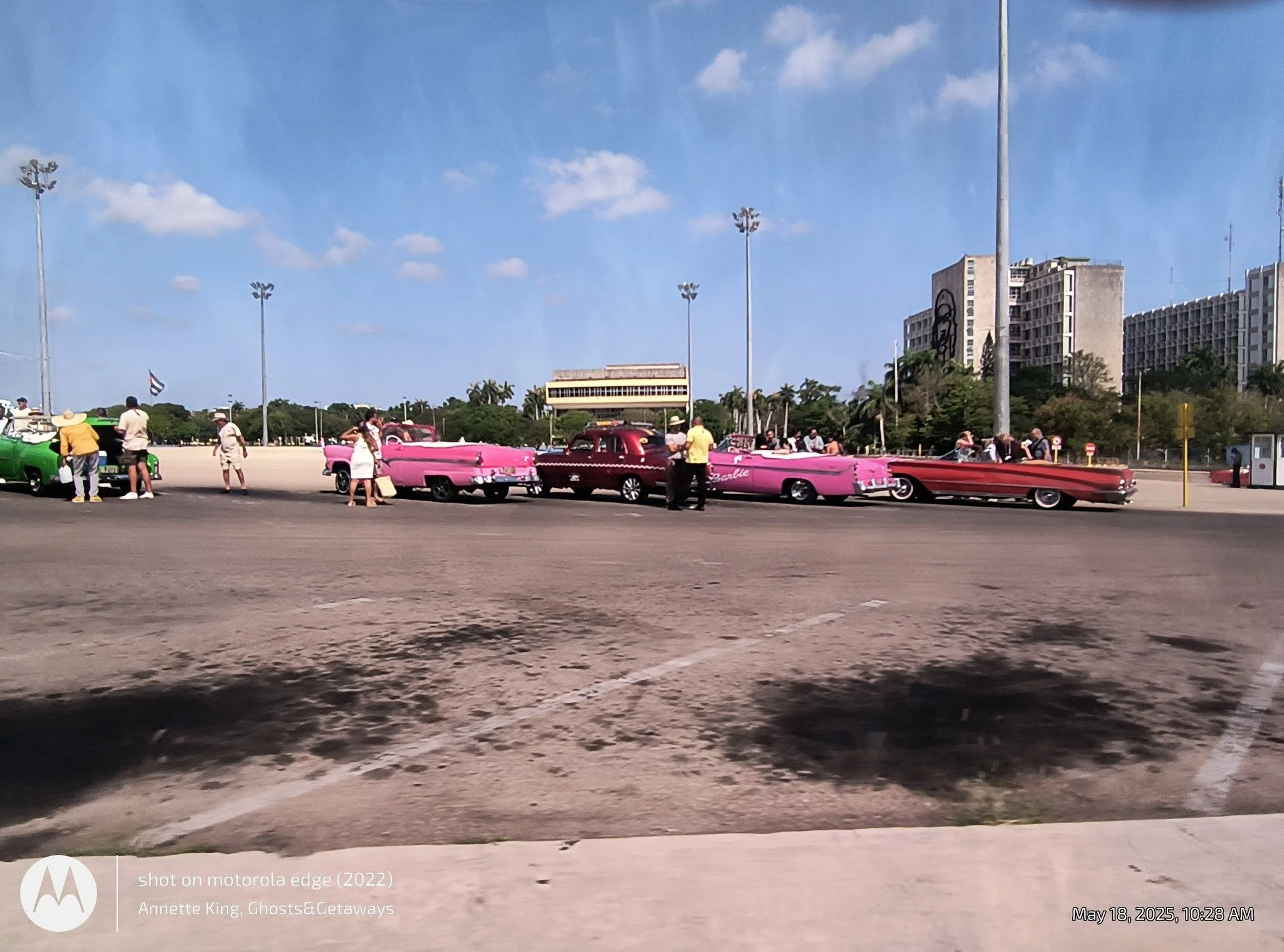 A scene at a parking lot with vintage pink and green cars on display, people walking around, and a clear blue sky with a few clouds.