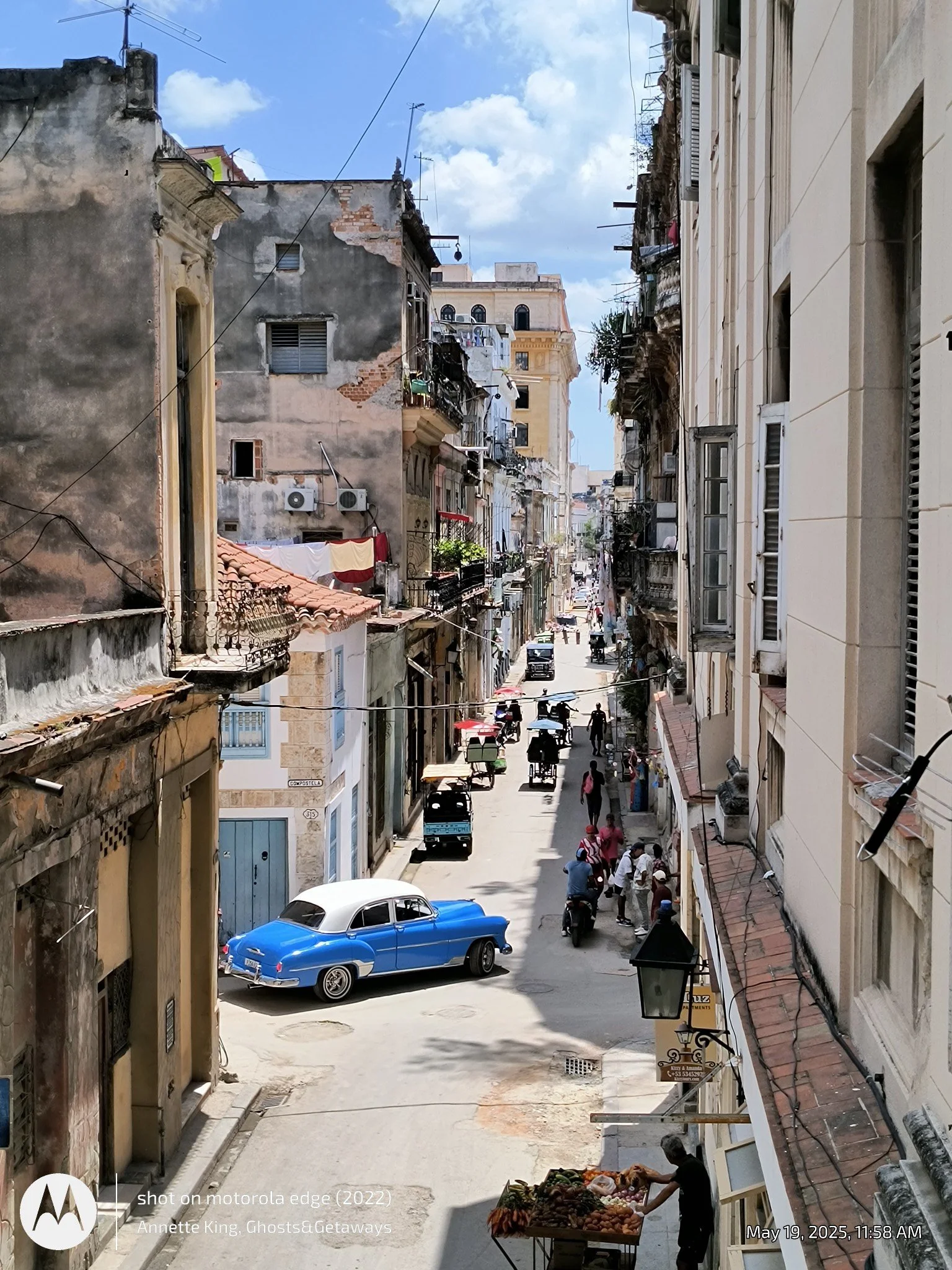 A narrow city street with old, weathered buildings on either side. The street has cars and pedestrians, with some vendors selling goods on the sidewalk. The sky is partly cloudy.