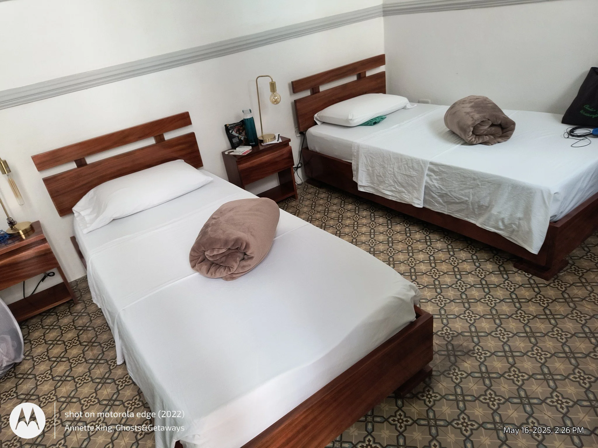 Two neatly made beds with white sheets, each with a rolled beige blanket at the foot, in a hotel room. A wooden nightstand between the beds holds a lamp, a bottle of water, and some personal items. The room has patterned tile flooring and white walls