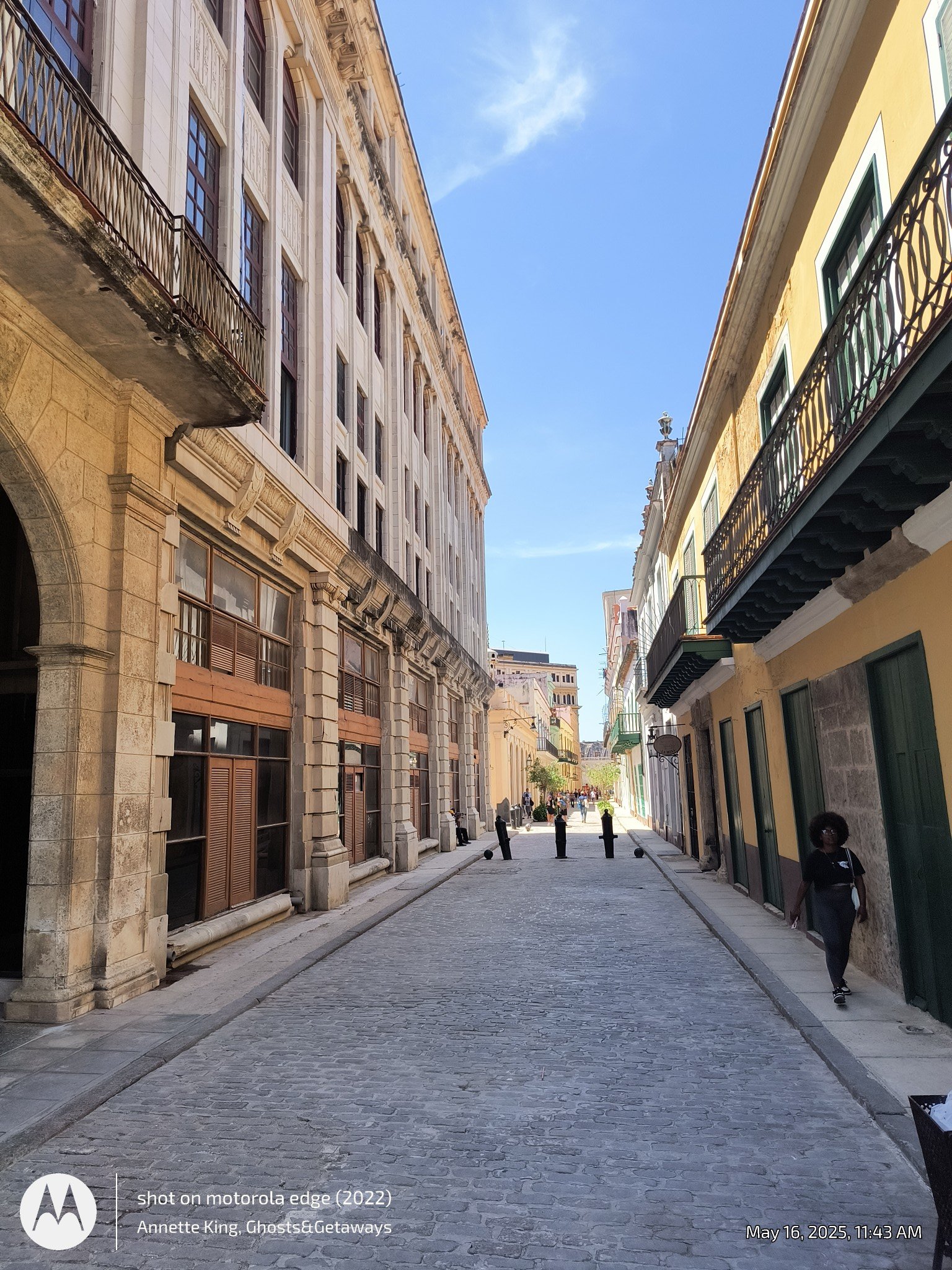 A narrow cobblestone street lined with historic multi-story buildings under a blue sky with some clouds. People are walking along the sidewalk and street.