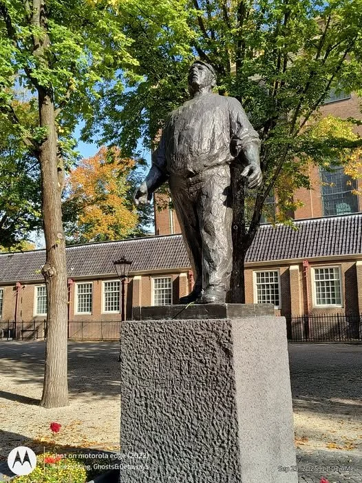 A bronze statue of a man standing on a stone pedestal outdoors, surrounded by trees and a brick building in the background, under a sunny sky.