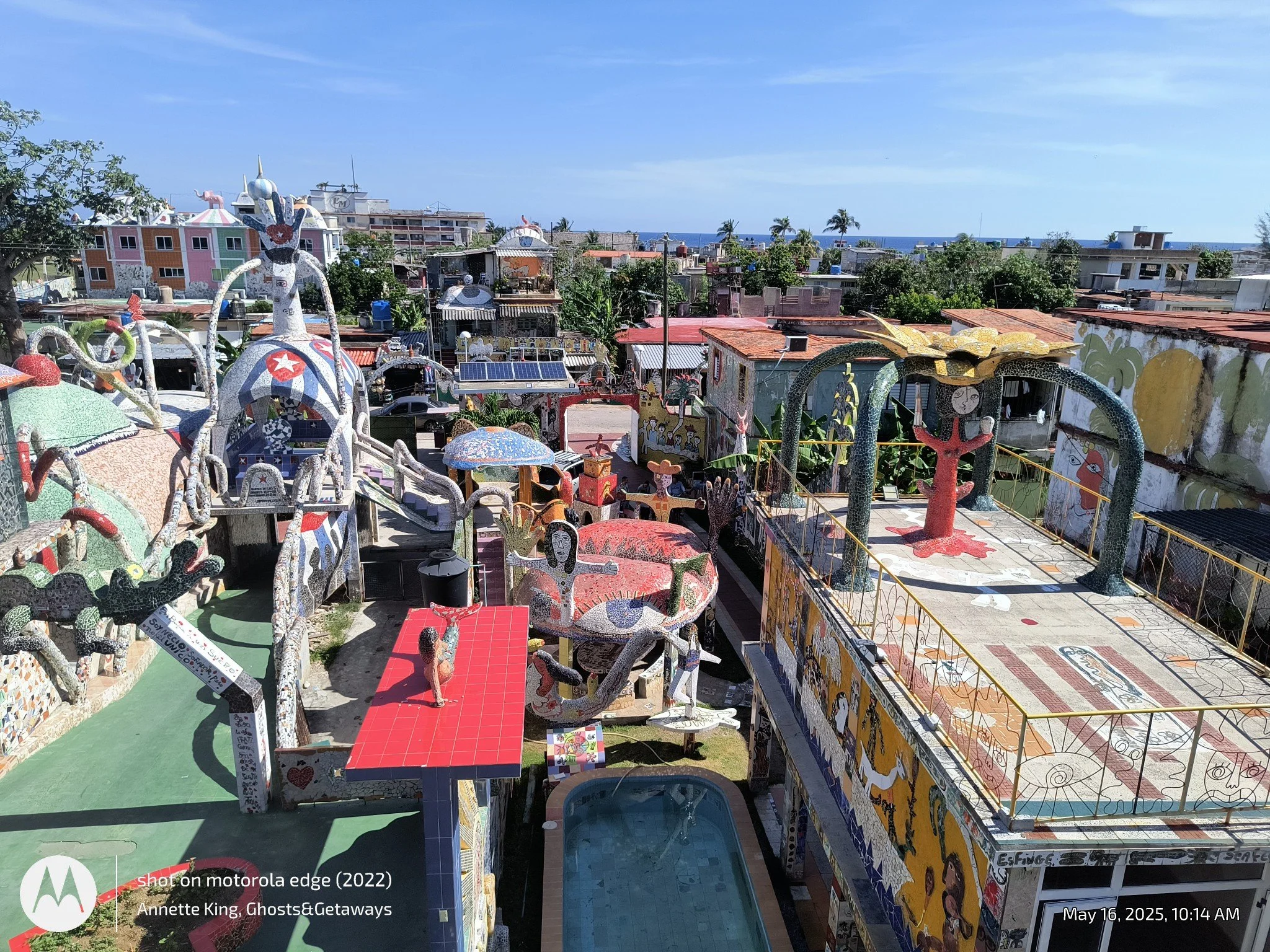 Colorful outdoor art installation with sculptures, mosaics, and painted walls in a vibrant coastal town with the ocean in the background.