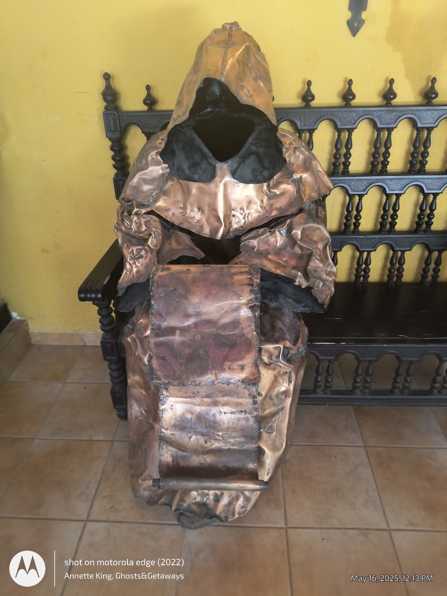 A metallic sculpture resembling a figure with a large, irregular head and an open face, sitting on a dark bench against a yellow wall.