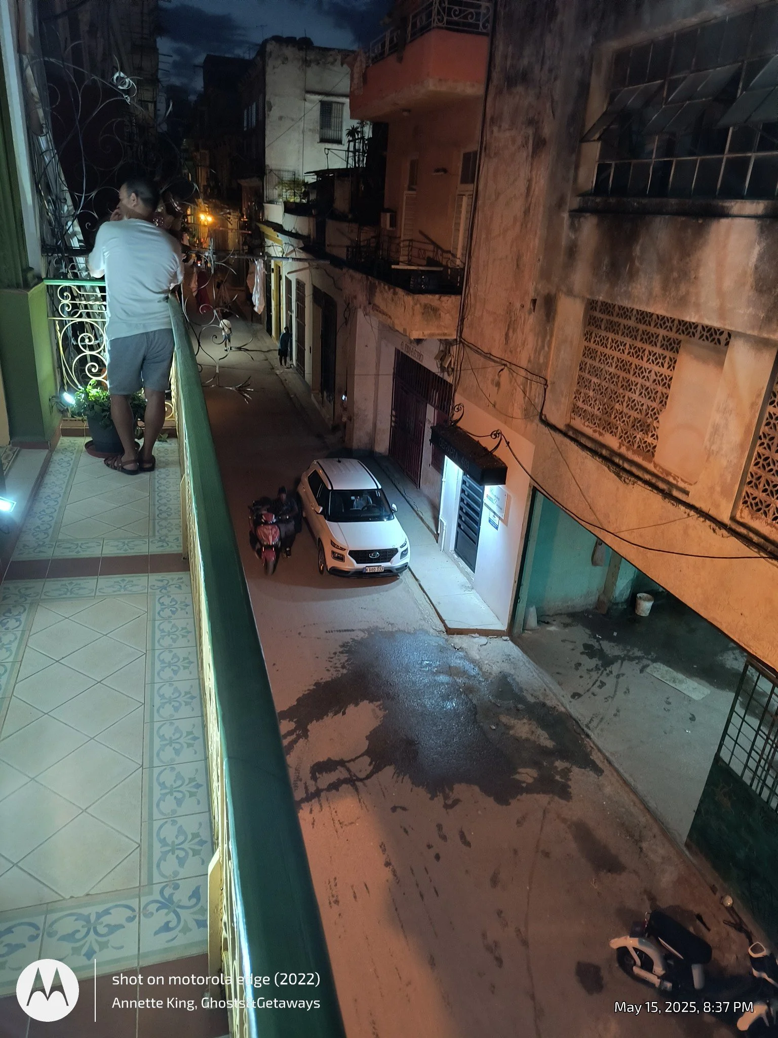 Nighttime street view with parked cars and motorcycles, a person standing on a balcony looking at the street, buildings with windows and balconies, and wet pavement reflecting streetlights.
