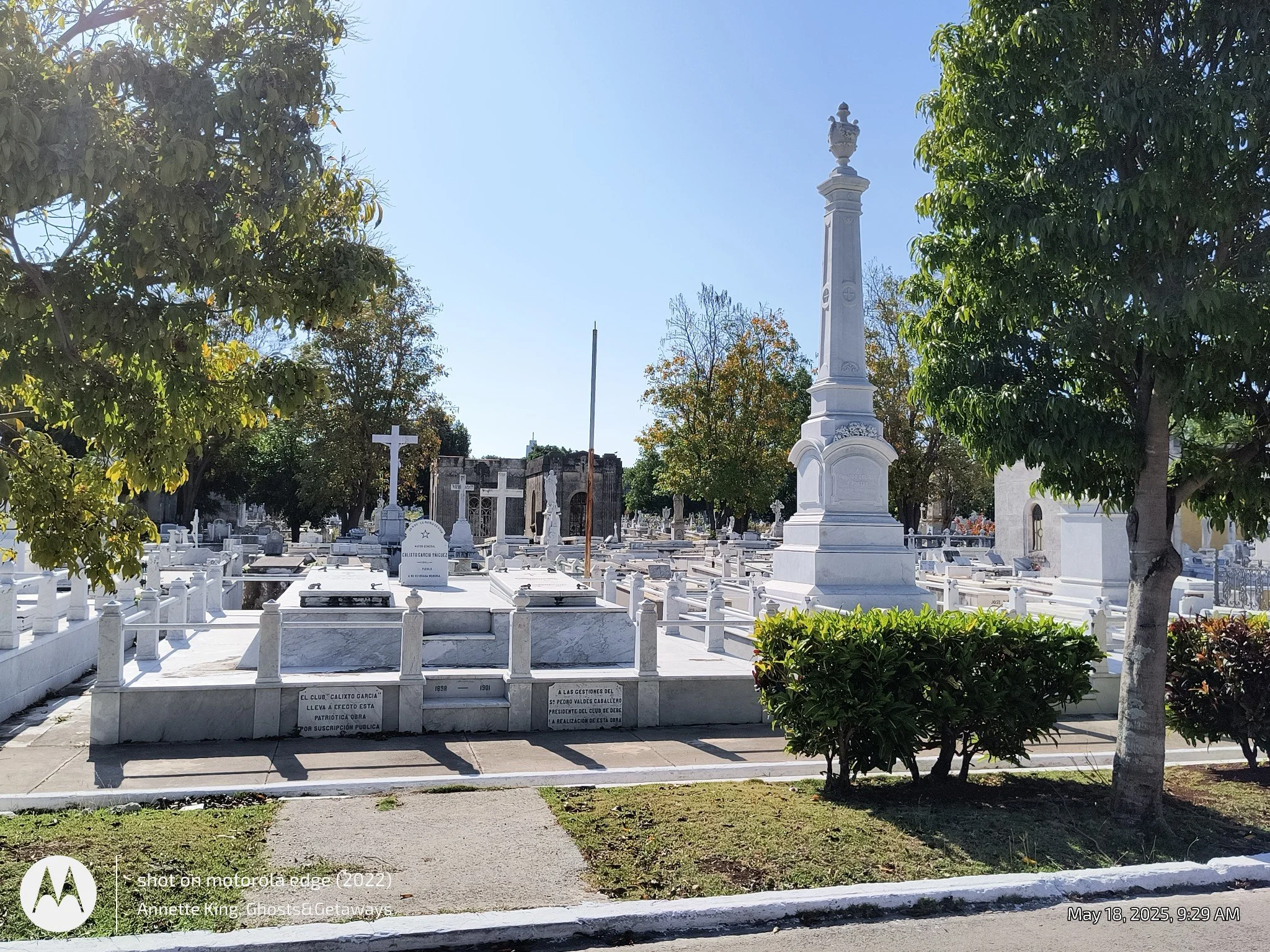 View of a cemetery with white marble graves and tombstones, a large white monument with a decorative urn on top, surrounded by green trees and bushes, under a clear blue sky.