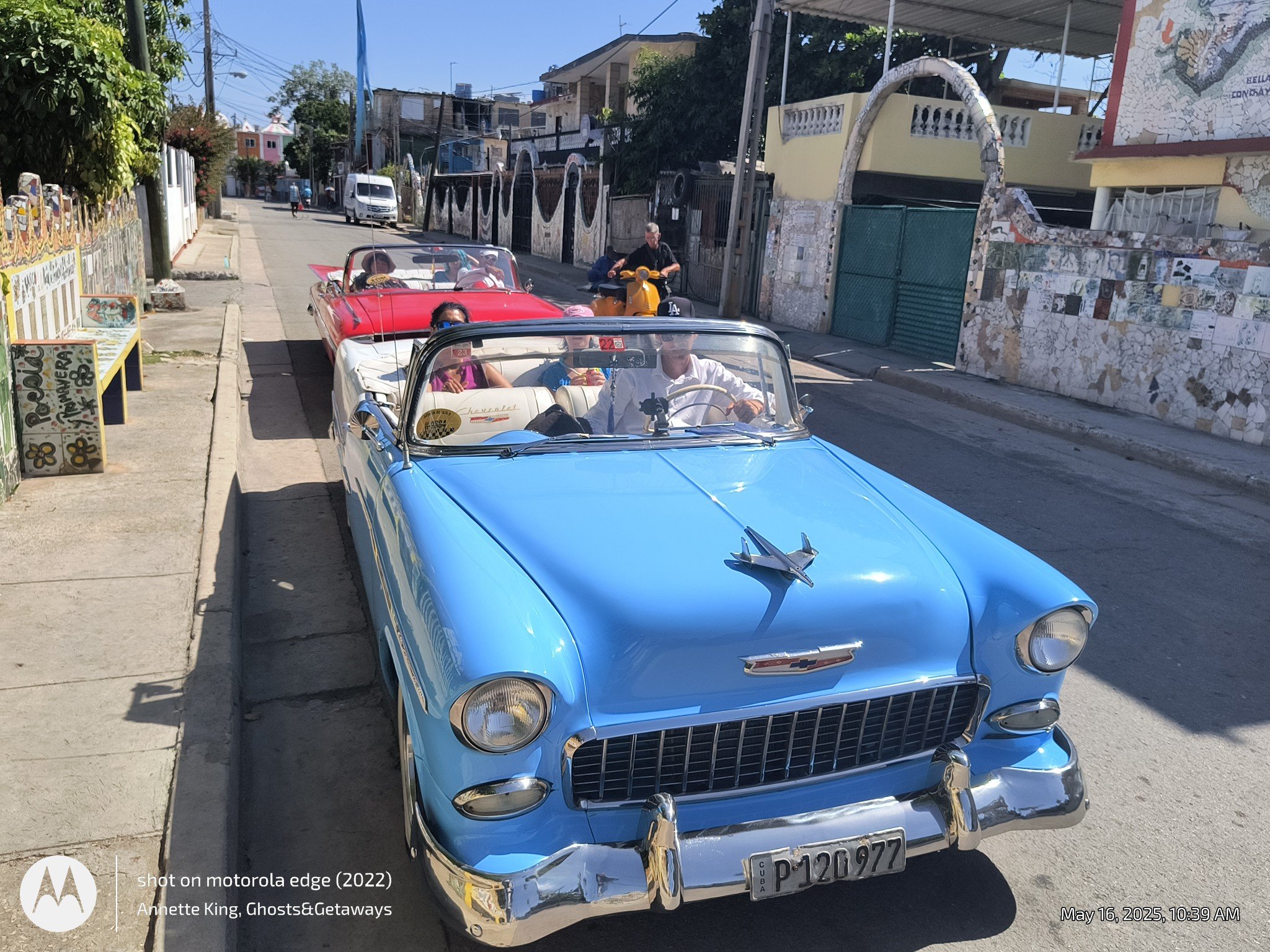 A street scene with a vintage blue Chevrolet convertible car in the foreground and a red convertible behind it, both cars with people inside. A person is riding a yellow scooter on the street. There are houses and trees lining the street, along with 